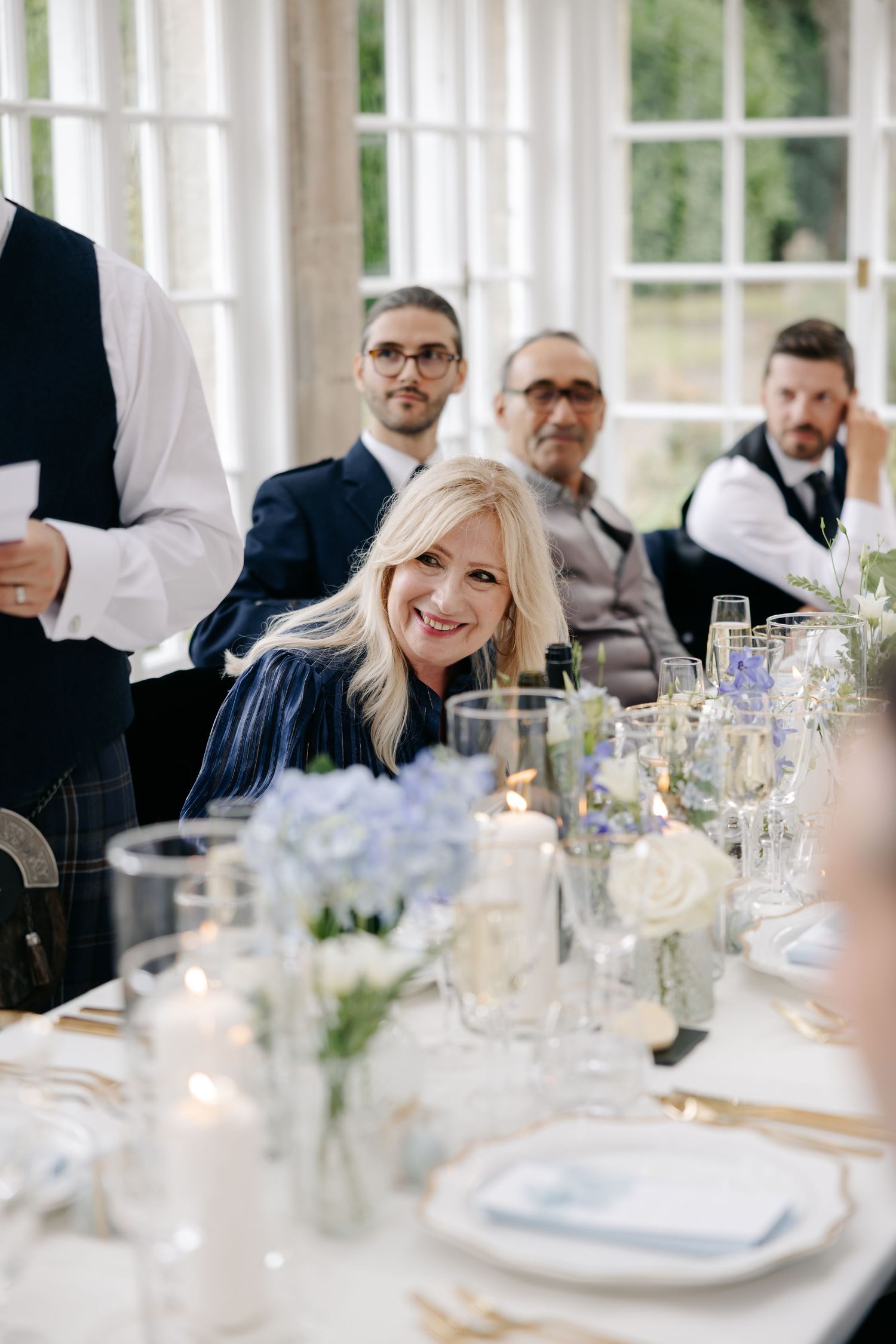 Smiling blonde woman at elegantly set wedding reception table with blue hydrangeas and candles.