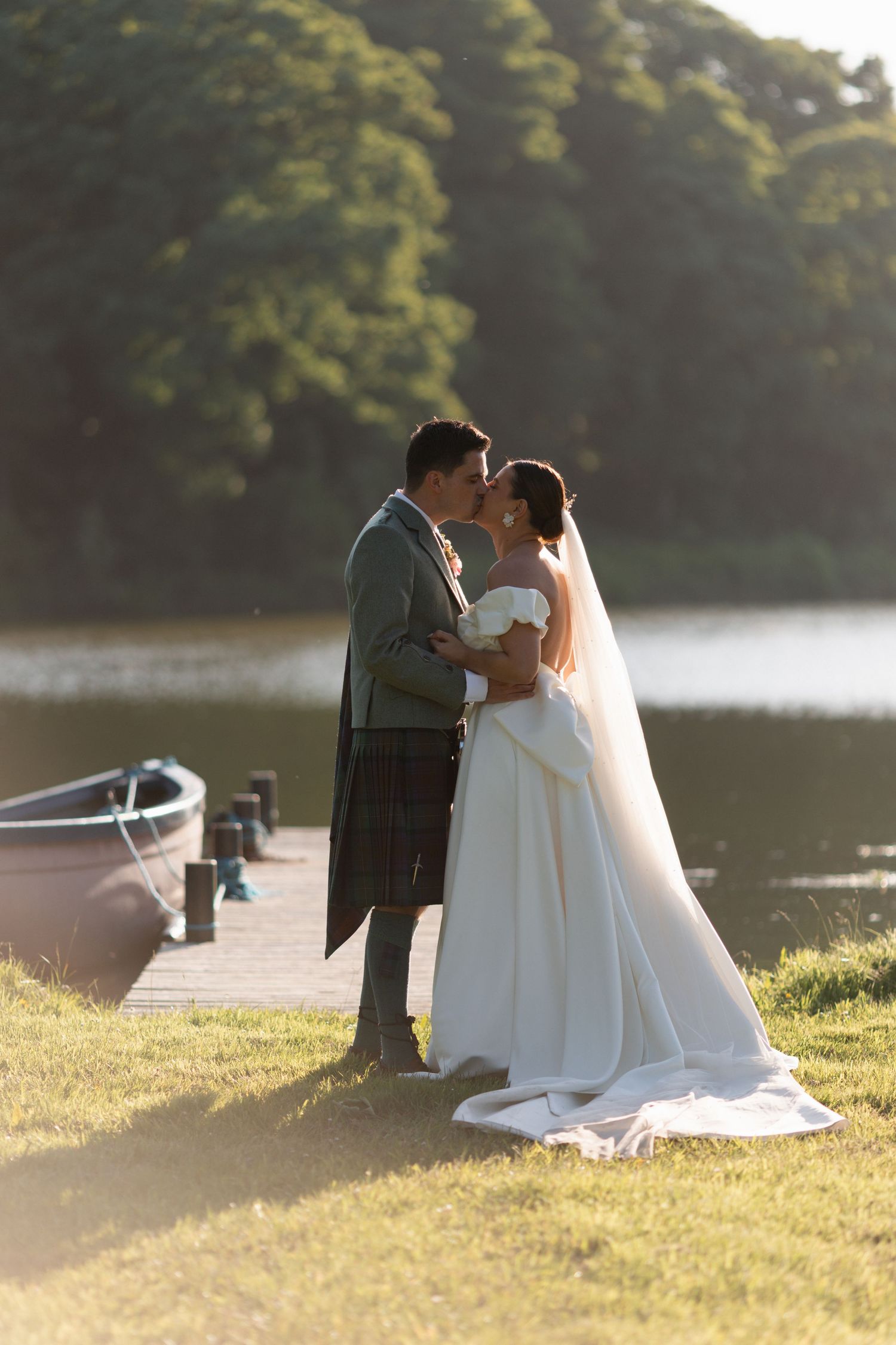 Bride and groom kissing by a lakeside dock, groom in Scottish kilt, bride in white gown with veil, golden hour light.