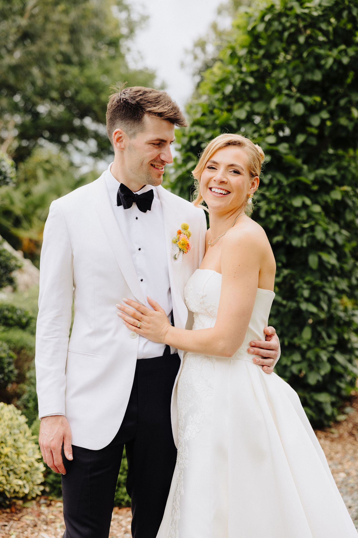 Smiling bride in strapless white gown embraces groom in white tuxedo jacket and bow tie outdoors.