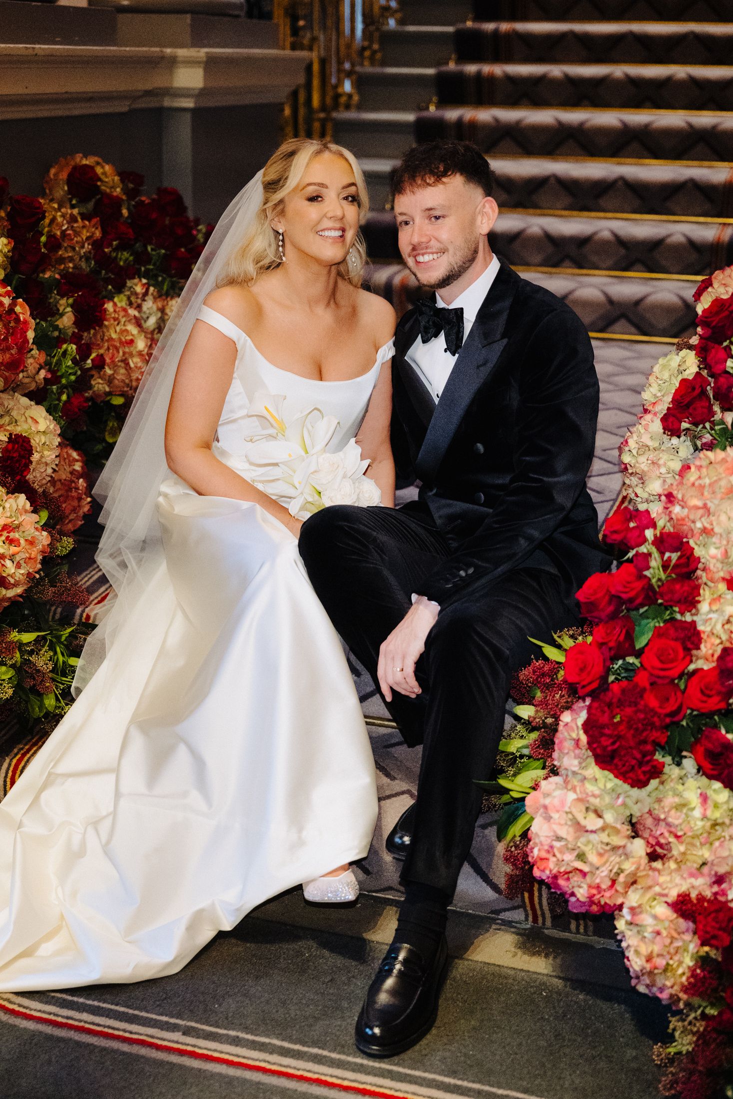 Bride in white gown and groom in black tuxedo sitting together surrounded by red and pink floral arrangements.