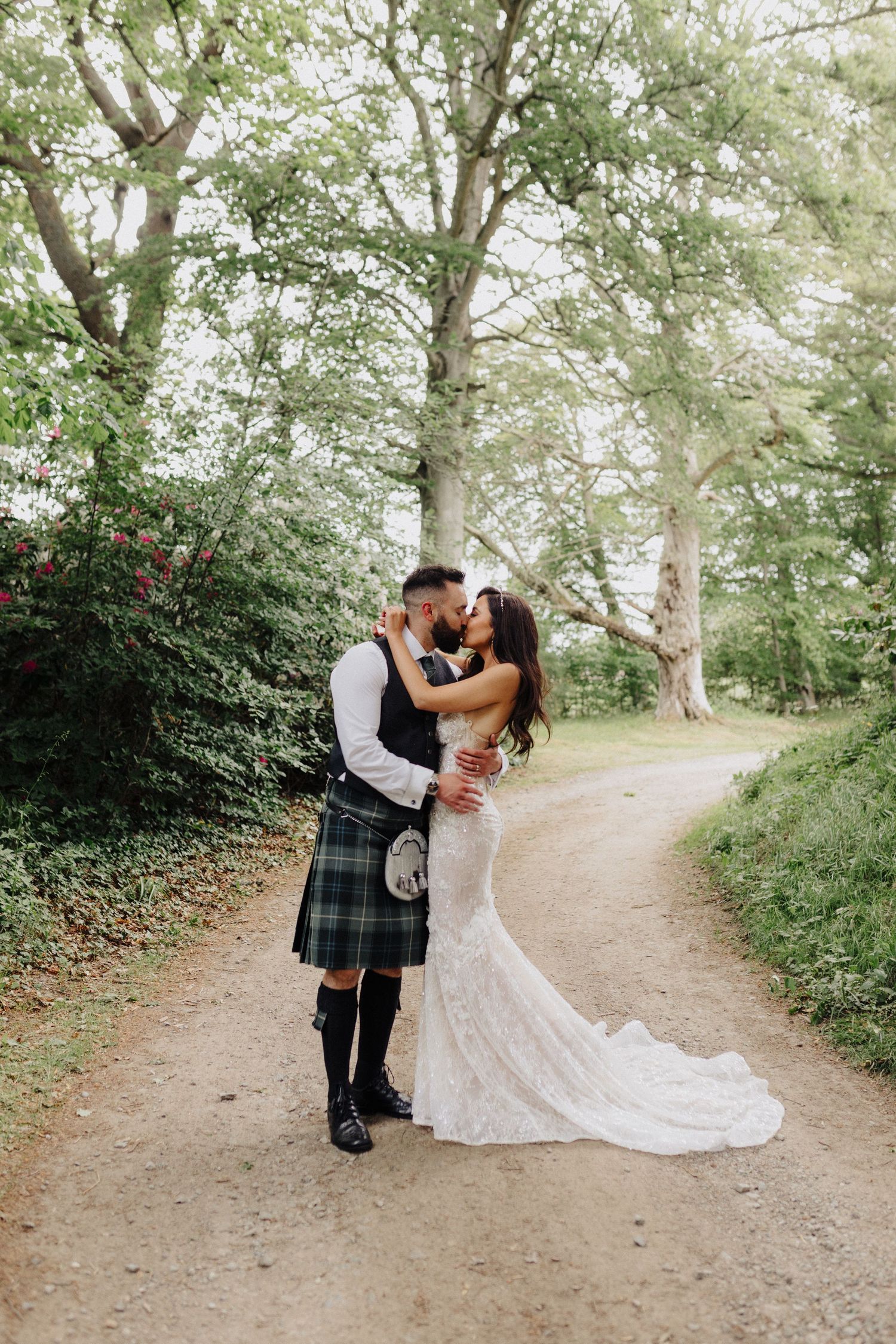 Bride and groom kissing on a woodland path, groom wearing a green tartan kilt, bride in a lace mermaid wedding gown.