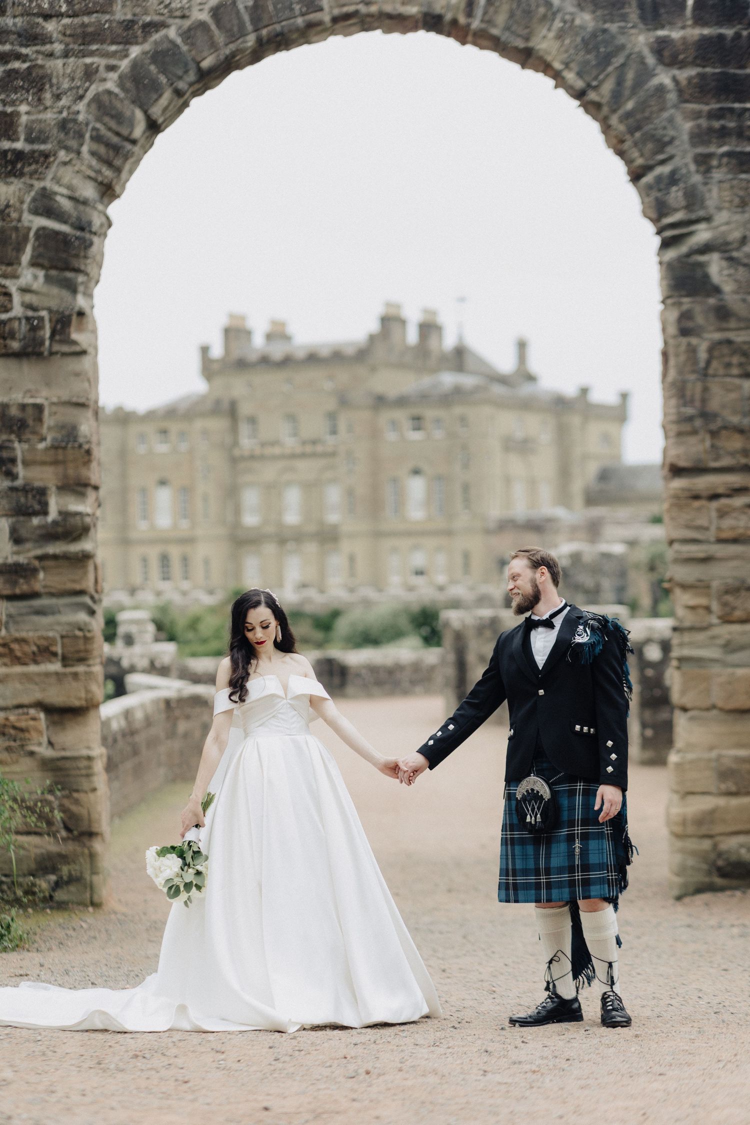 Bride in white gown and groom in Scottish kilt hold hands under stone arch with castle backdrop.