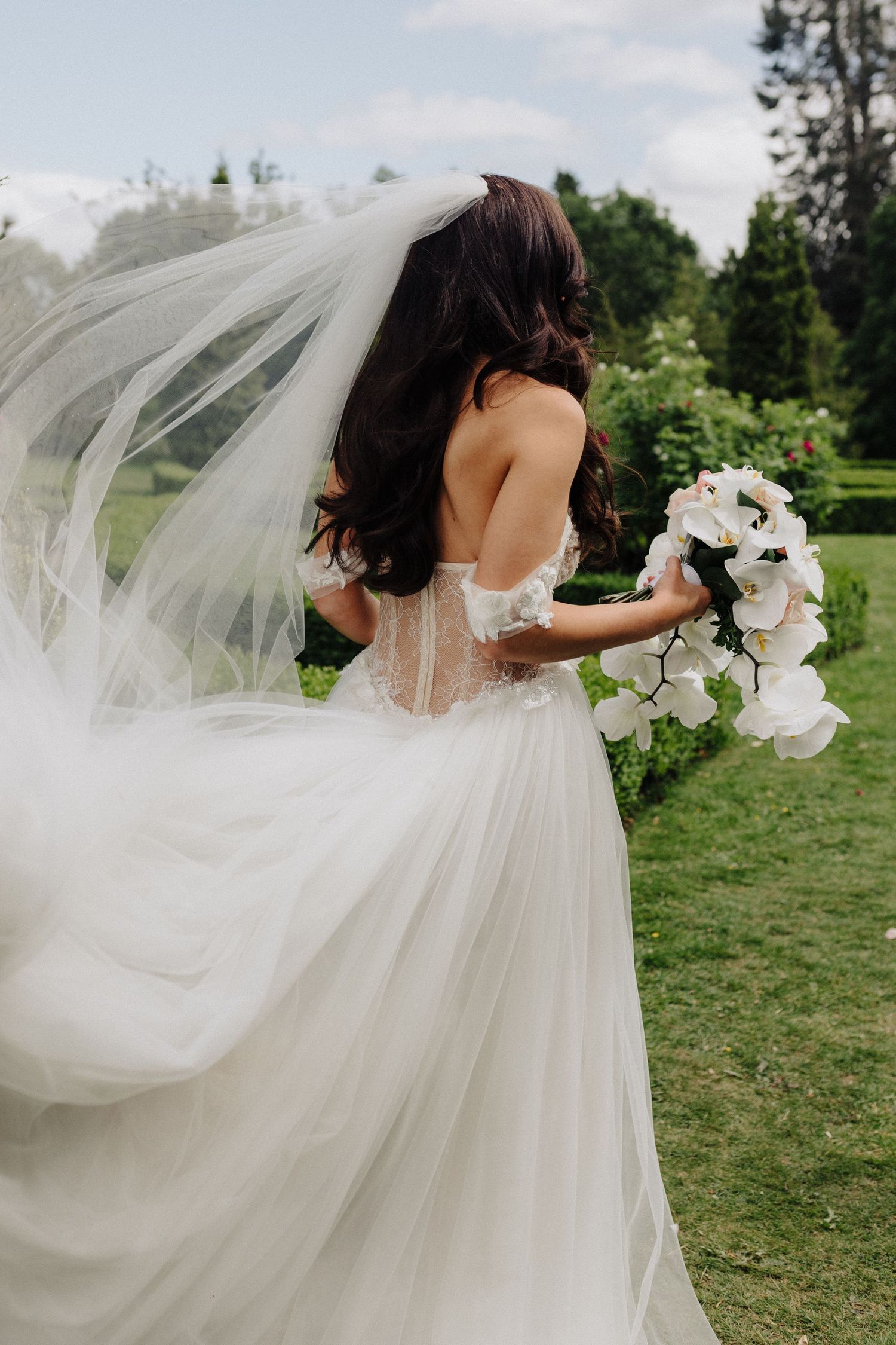 Bride in flowing white tulle gown with corset back holds white orchid bouquet in a lush garden.