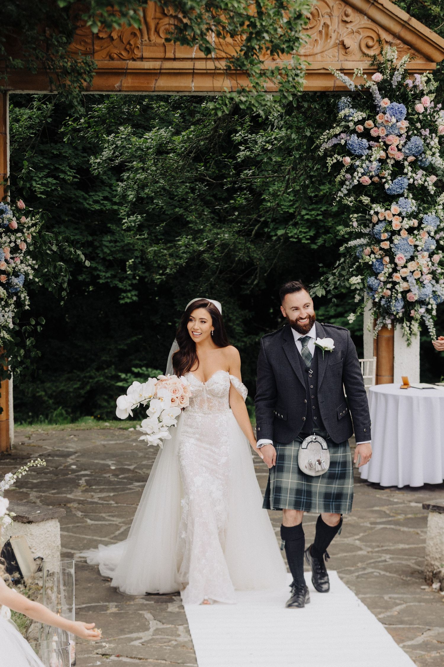 Bride in lace gown and groom in Scottish kilt walking the aisle at an outdoor floral ceremony.