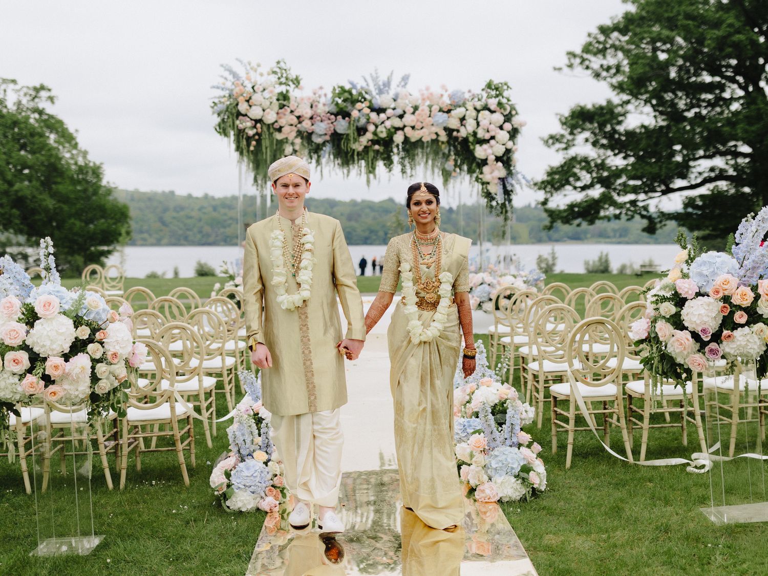 Bride and groom in gold traditional attire hold hands at an outdoor wedding ceremony with floral arch backdrop.