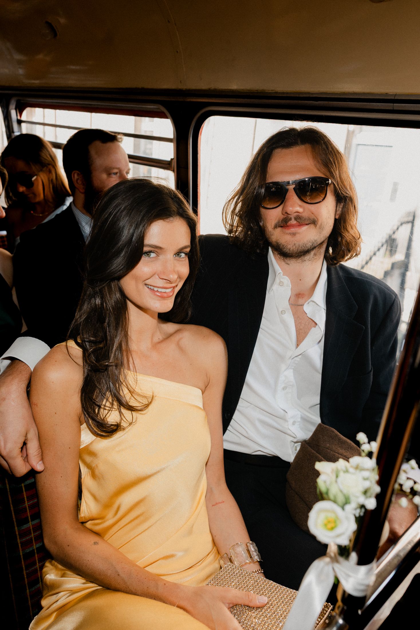 A couple in formal attire smiles while seated inside a vintage bus, woman in yellow dress, man in sunglasses.