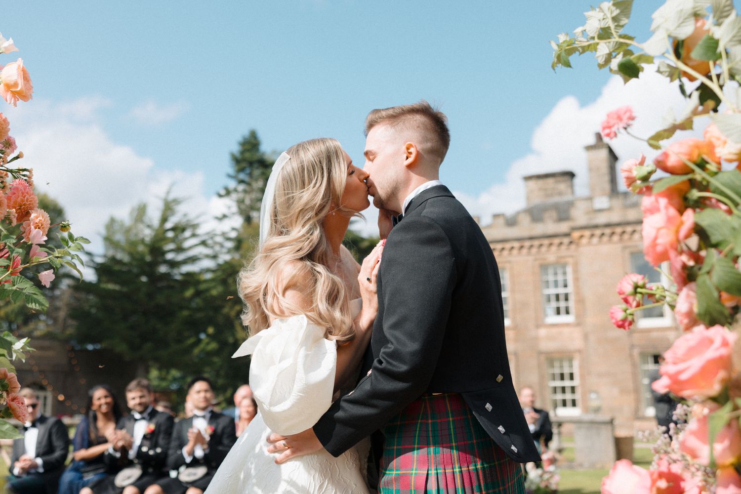Bride and groom share first kiss at outdoor Scottish wedding ceremony, groom wearing traditional tartan kilt.