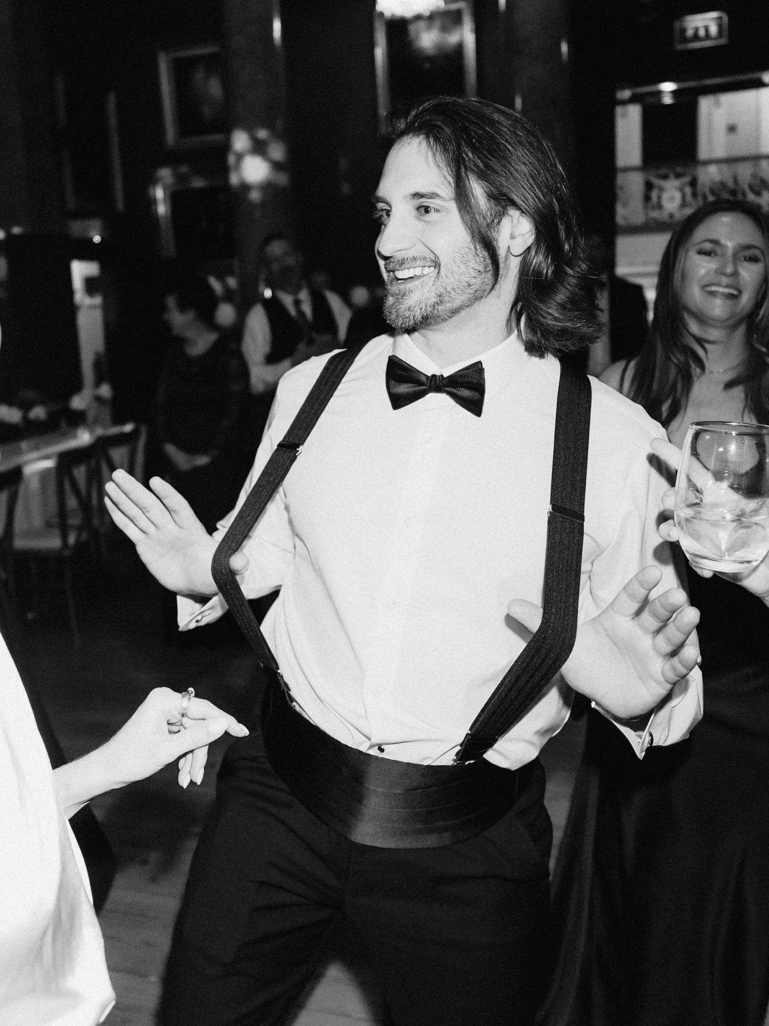 Groom dancing joyfully in black and white photo, wearing suspenders and bow tie at wedding reception.