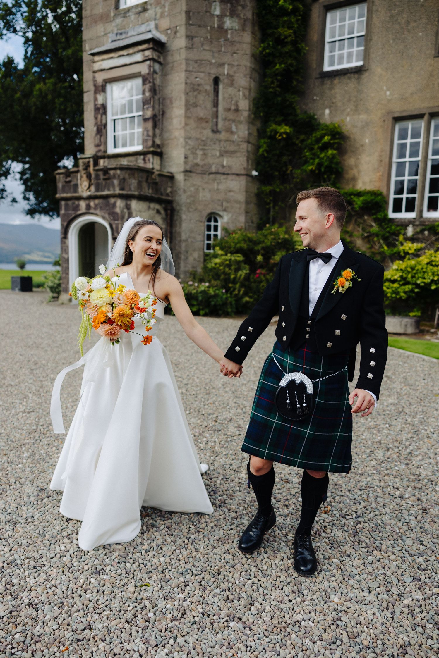 Laughing bride in white gown and groom in green tartan kilt hold hands outside a Scottish estate.