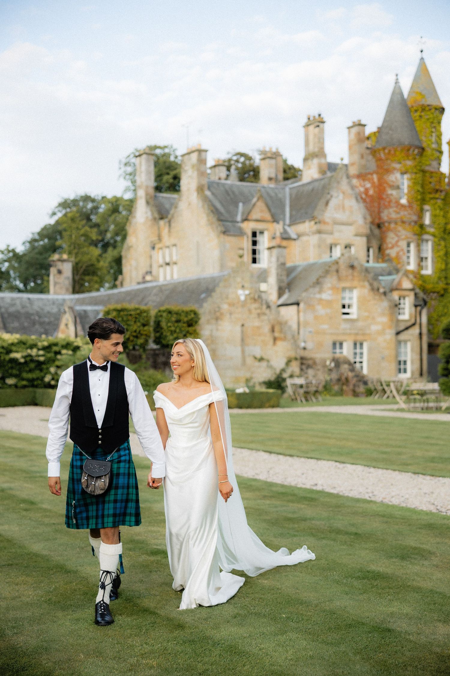 Groom in tartan kilt and bride in off-shoulder gown walk hand-in-hand before a Scottish castle.