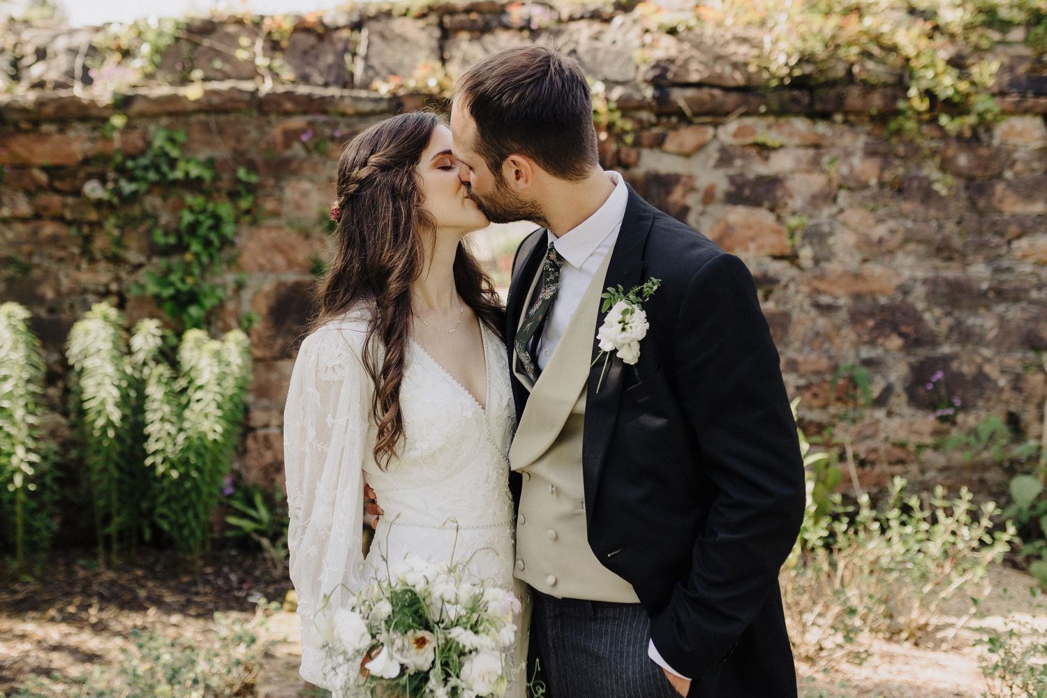 Bride and groom sharing a romantic kiss outdoors, bride holding white bouquet, groom in black suit with boutonniere.