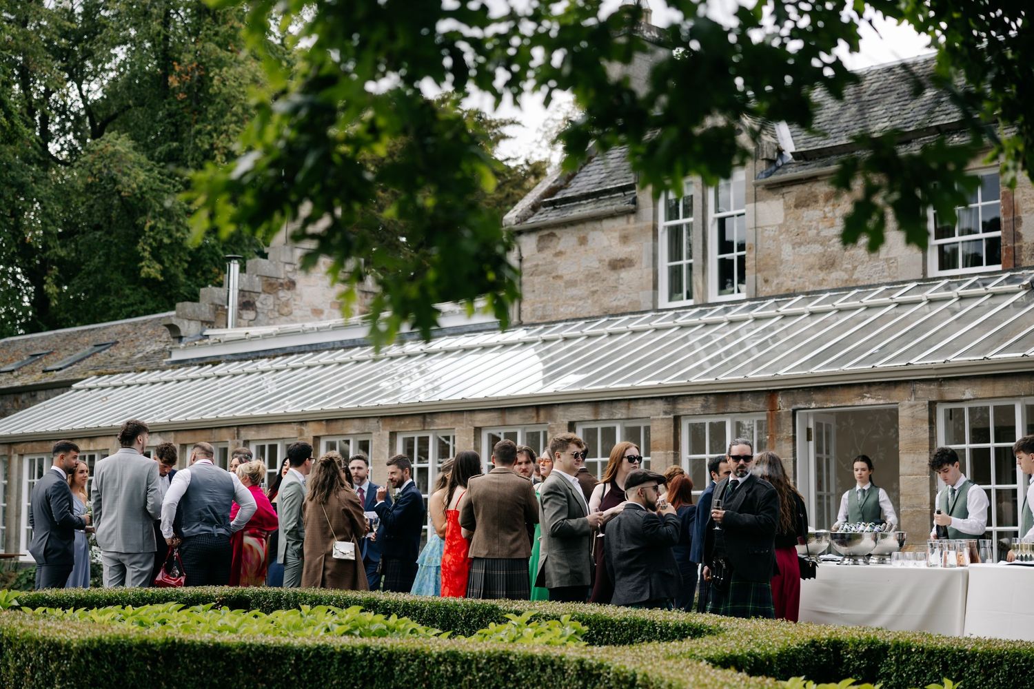 Elegant outdoor wedding reception at a stone manor house with guests mingling in manicured gardens.