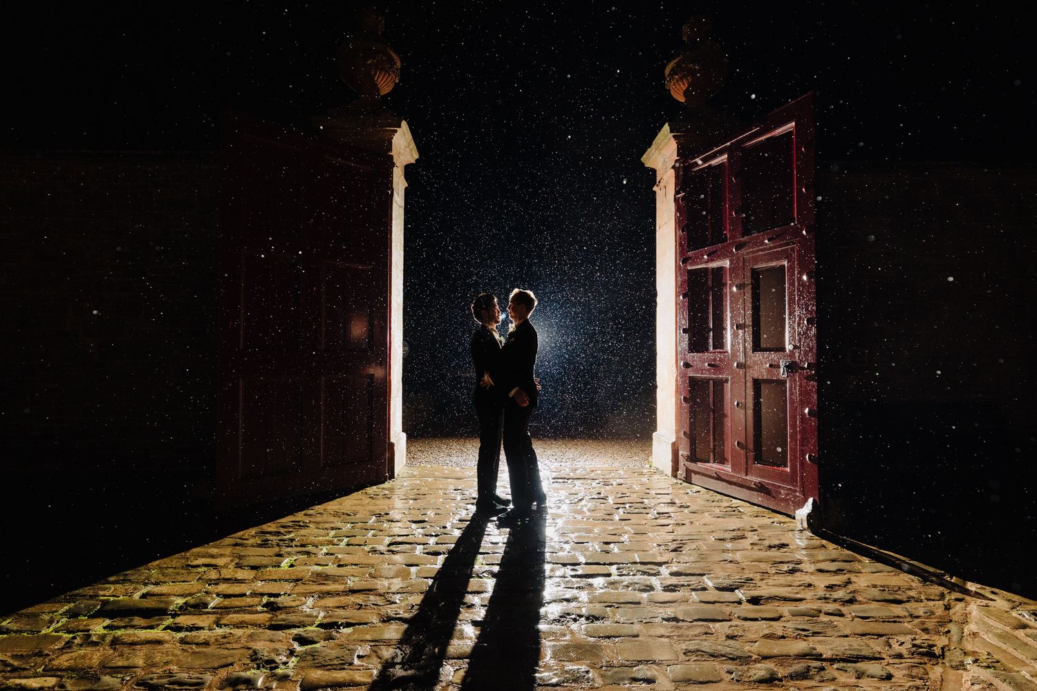 Couple embracing in rain at night between ornate red doors on cobblestone path with dramatic backlighting.