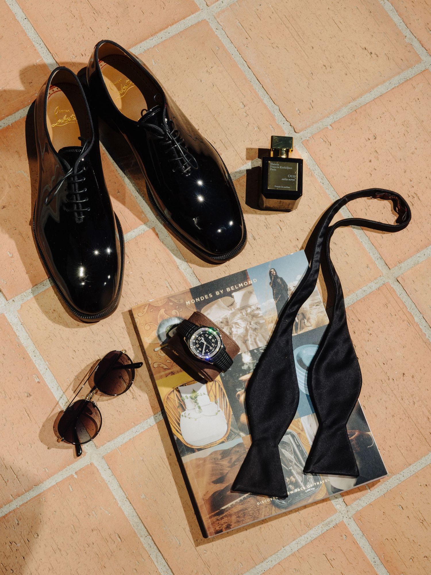 Flat lay of groom wedding accessories including black patent shoes, bow tie, watch, sunglasses, and cologne.