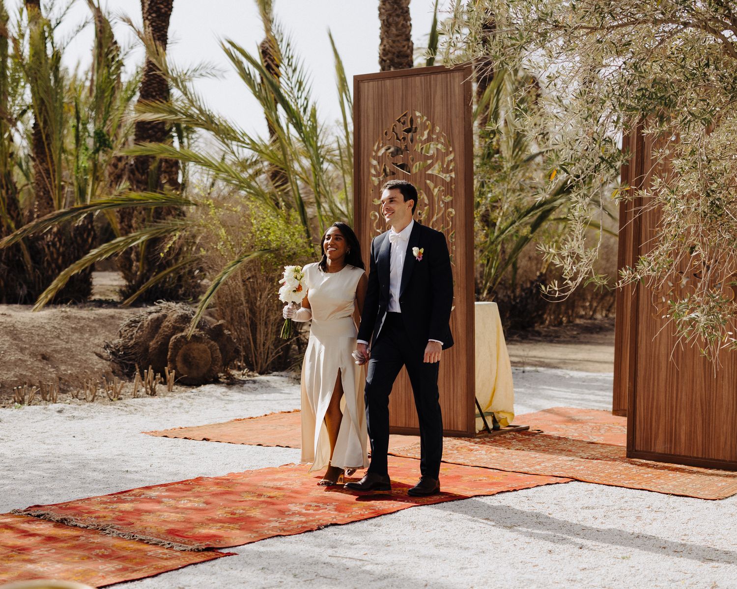 Bride and groom walk hand-in-hand at an outdoor desert wedding ceremony surrounded by palm trees and colorful rugs.