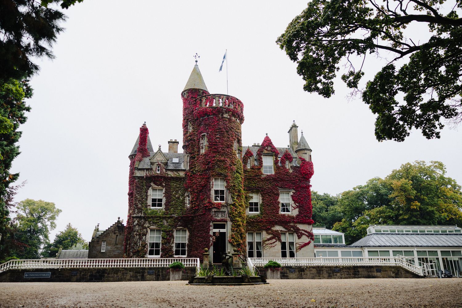 A historic Scottish castle covered in vibrant red ivy, featuring turrets and a flag, surrounded by gravel paths and trees.