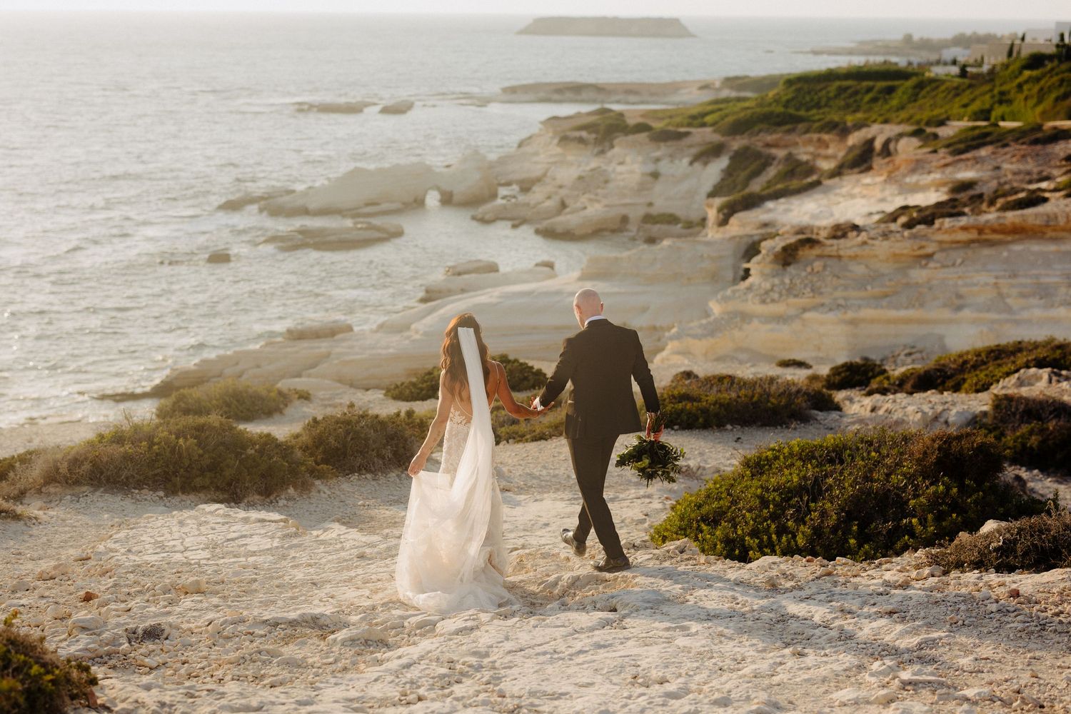 Bride and groom walking hand in hand along a rocky coastal shoreline during golden hour at their beach wedding.