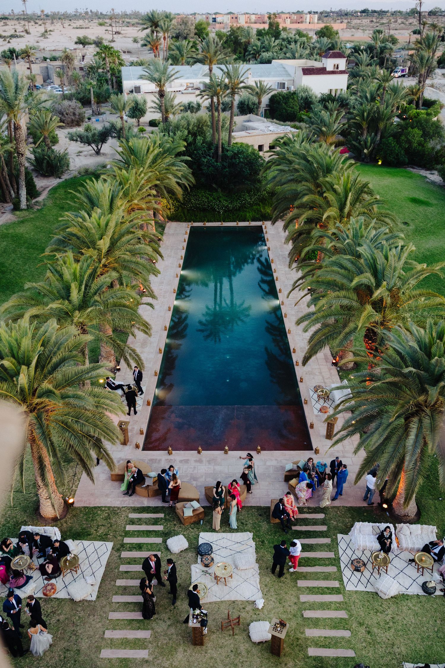 Aerial view of a Moroccan wedding venue with a long pool surrounded by palm trees and guests.