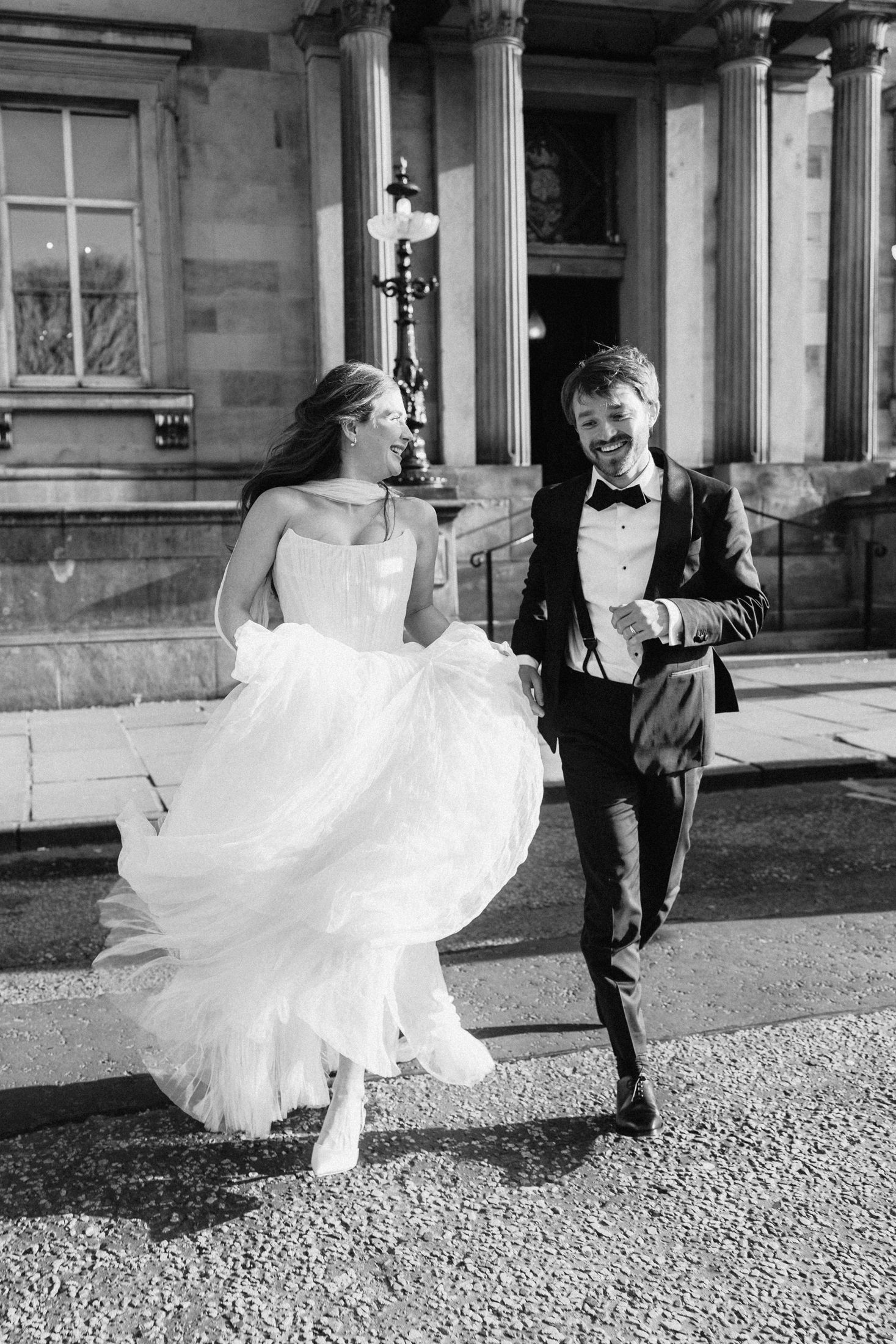 Black and white photo of laughing bride and groom running outside a grand stone building.