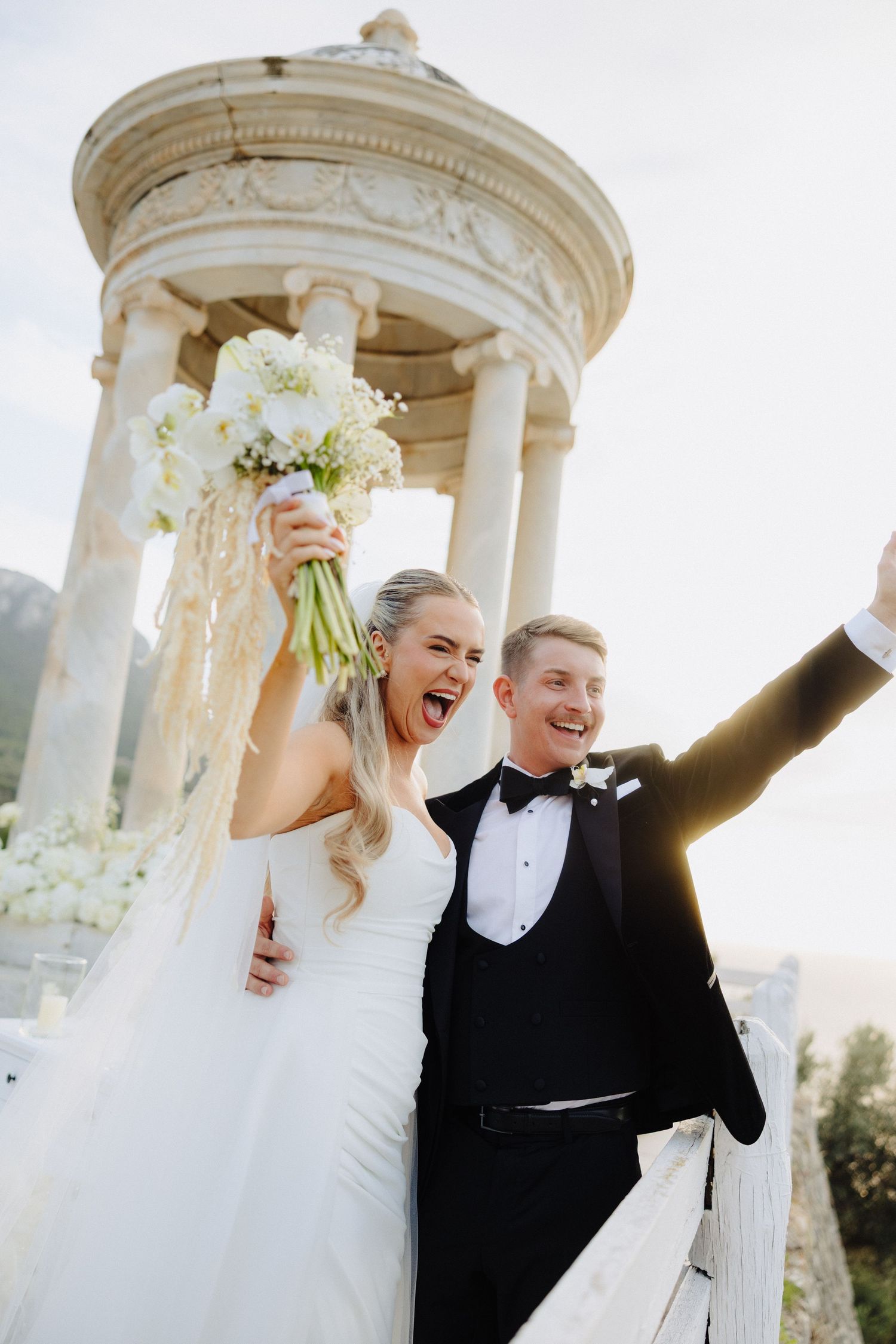 Joyful bride and groom celebrating at elegant white rotunda, bride raising bouquet, groom with arm raised in triumph.
