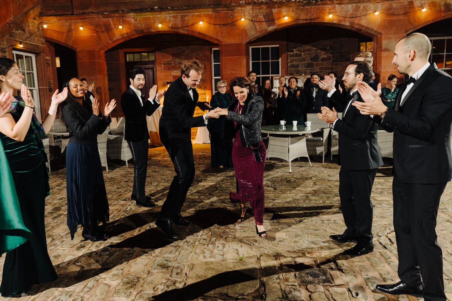Guests clapping and dancing at an elegant wedding reception in a brick venue with string lights.