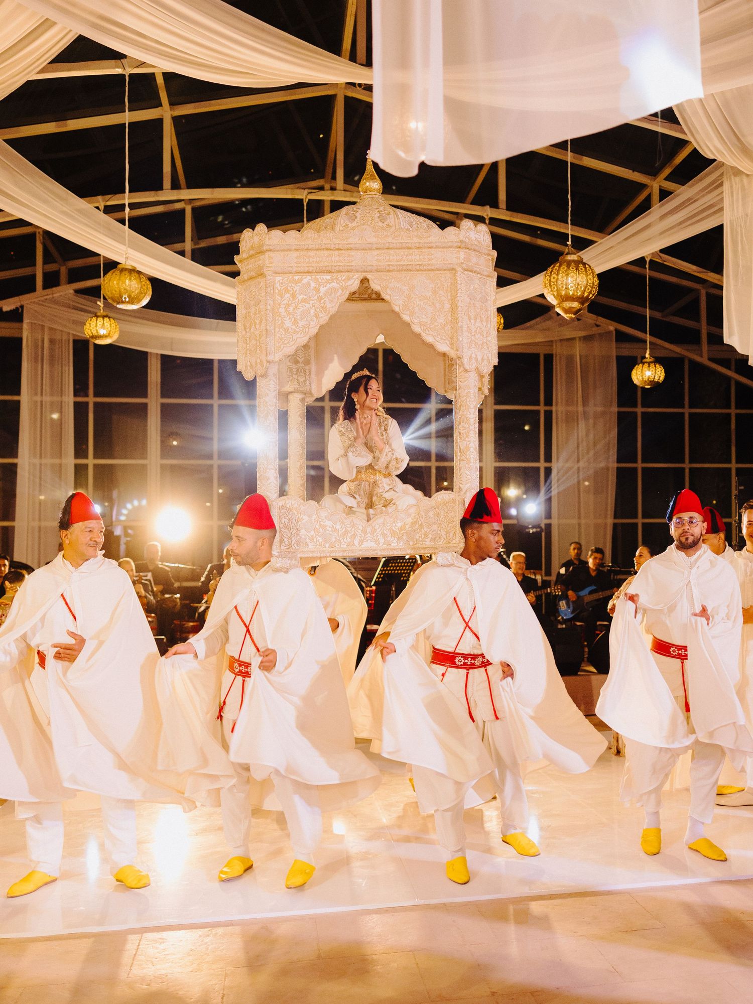 Bride carried in ornate white palanquin by men in traditional Moroccan white and red ceremonial attire.