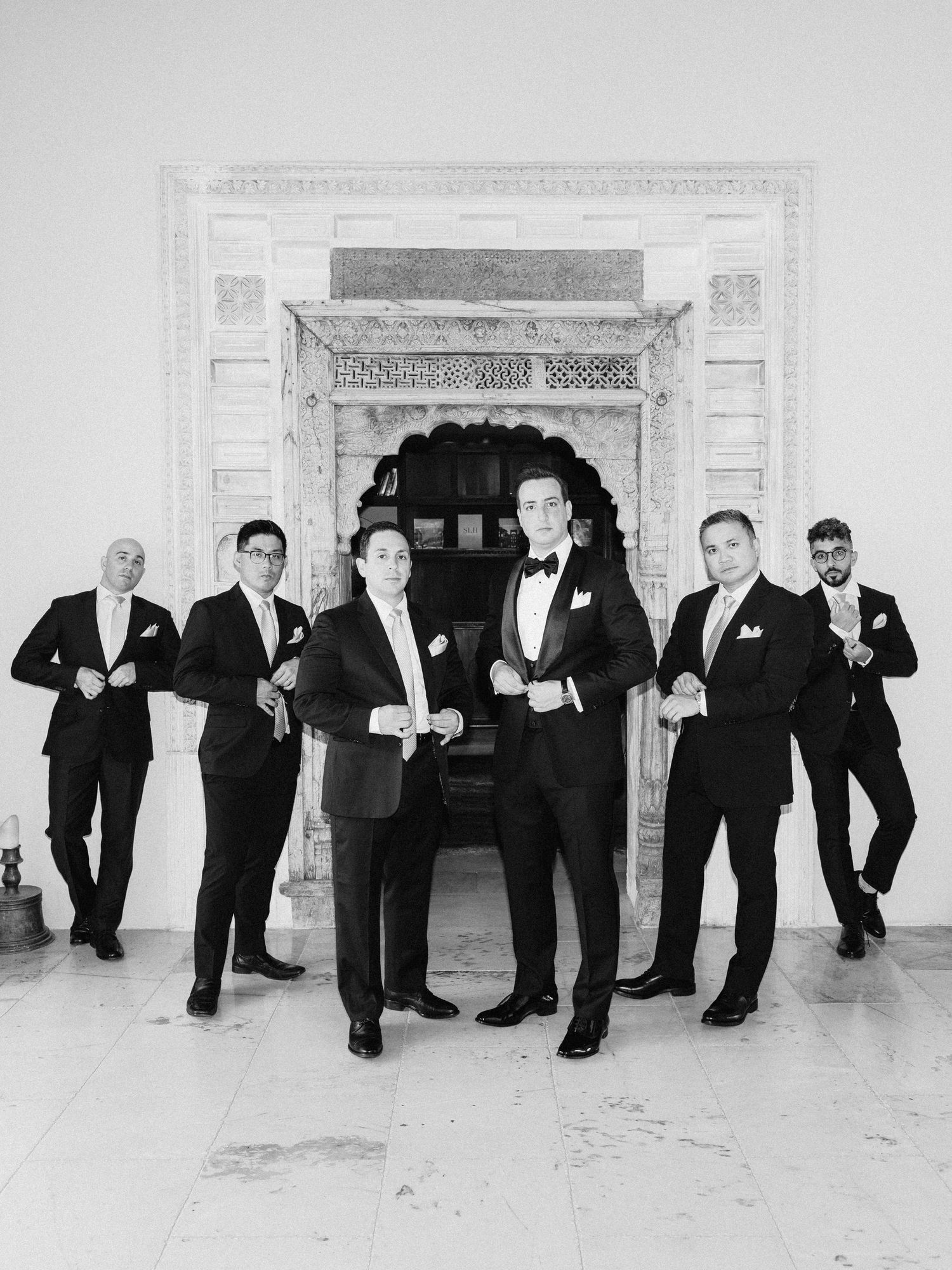 Black and white photo of a groom and groomsmen in tuxedos posing by an ornate fireplace.