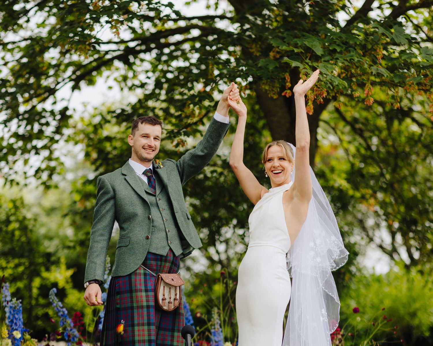 Joyful newlyweds celebrate outdoors, groom in Scottish tartan kilt and bride in white gown raising hands under lush green trees.