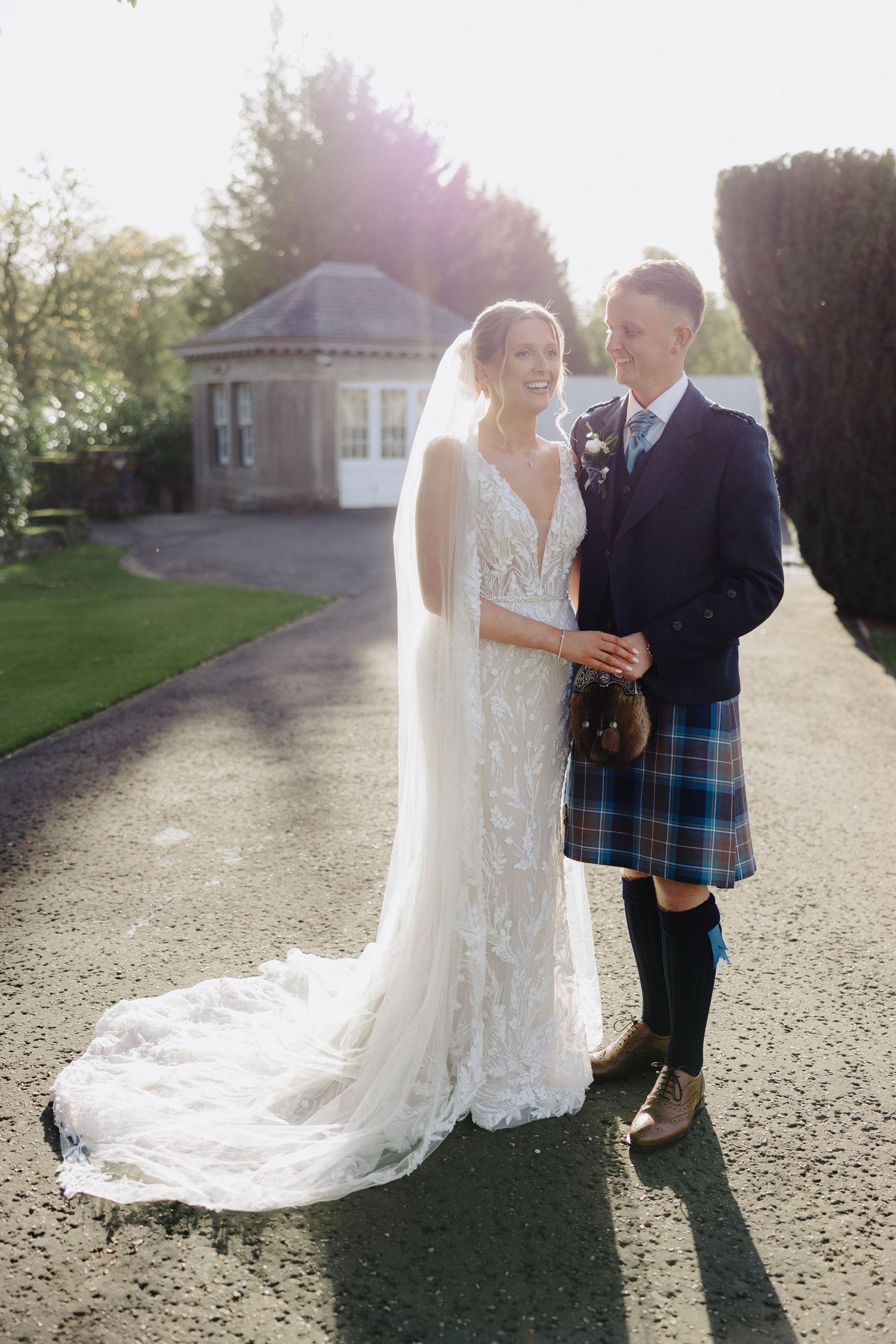 Bride in lace gown with veil and groom in blue tartan kilt hold hands outdoors in golden sunlight.