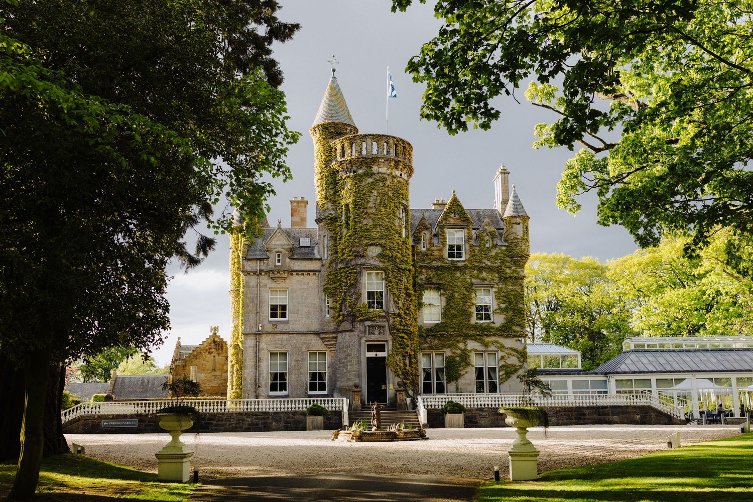 Ivy-covered Scottish castle hotel with turret, surrounded by lush trees and manicured grounds on a sunny day.