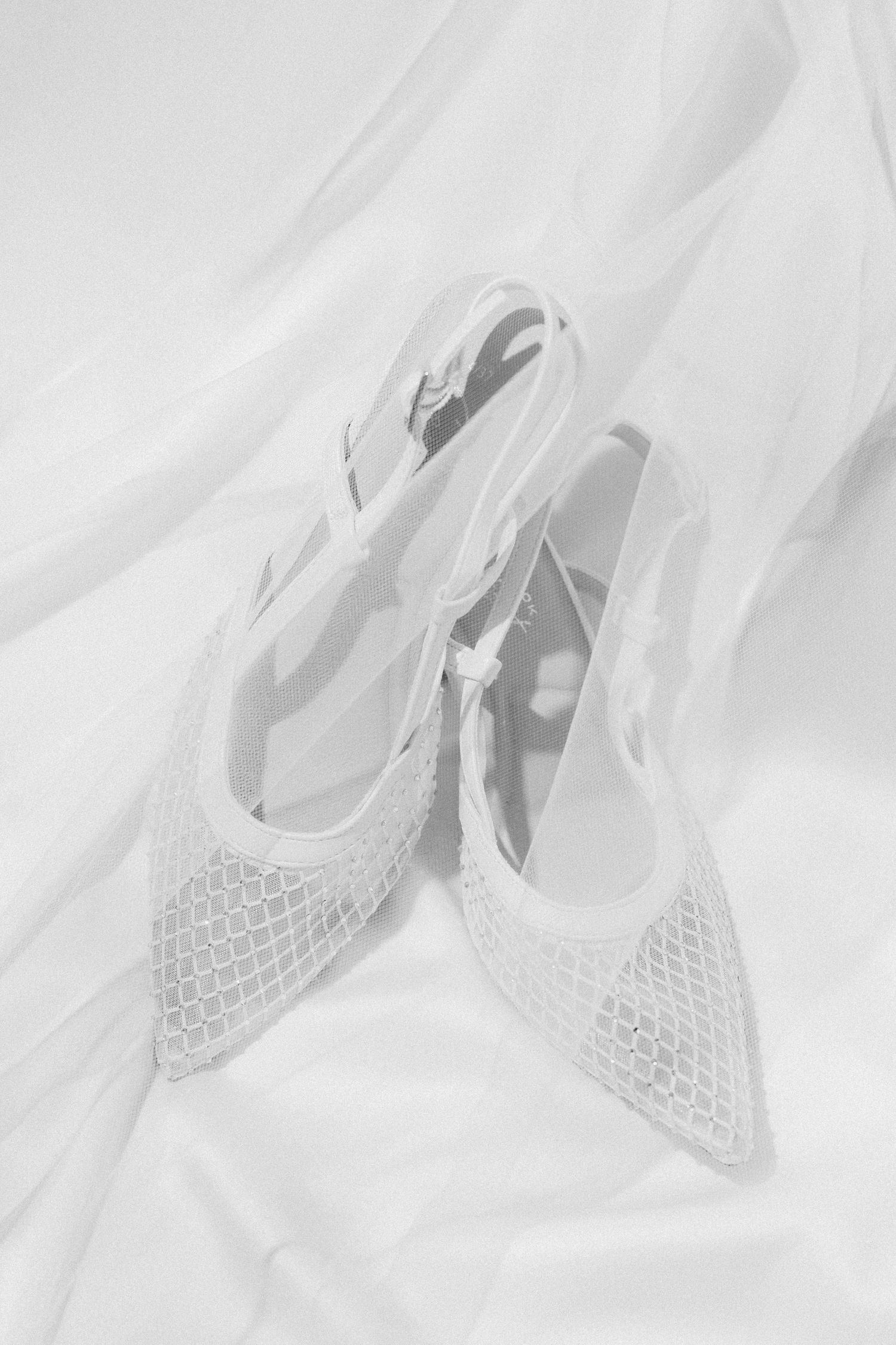 Delicate white mesh slingback bridal heels with pointed toe resting on soft white fabric backdrop.
