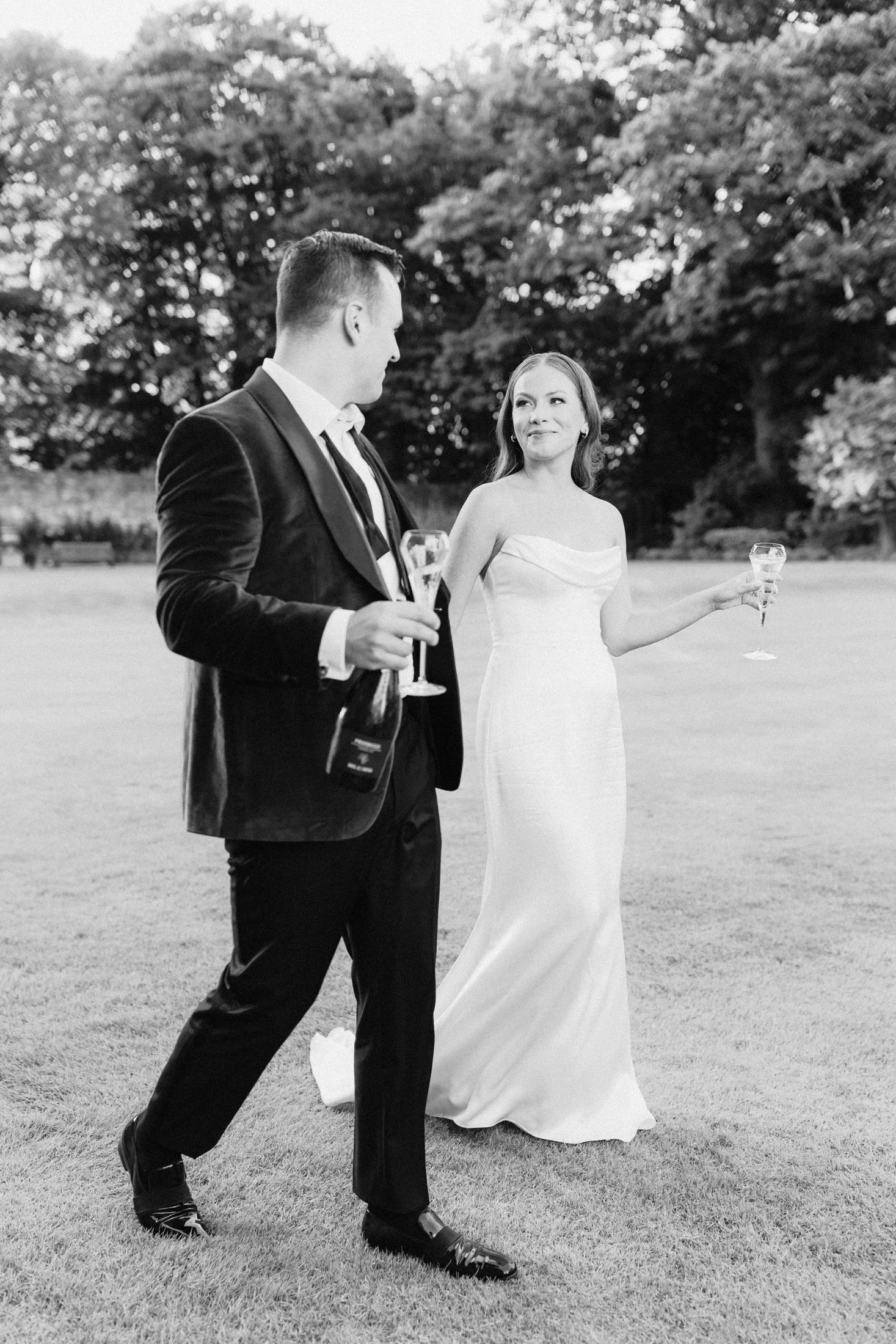 Black and white photo of bride and groom walking on lawn holding champagne glasses at elegant outdoor wedding.