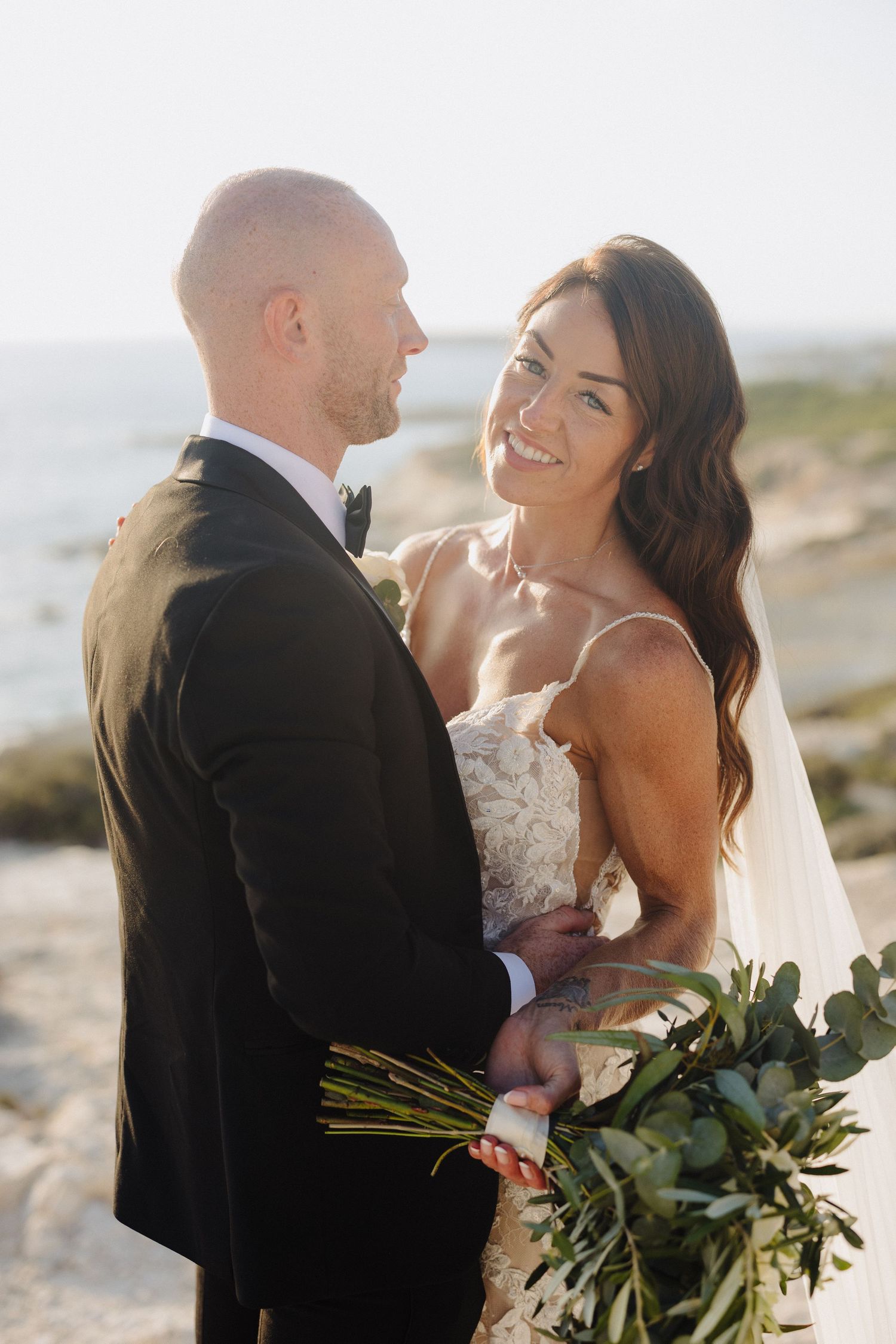 Bride in lace gown smiles at groom in black tuxedo holding greenery bouquet on rocky seaside at golden hour.
