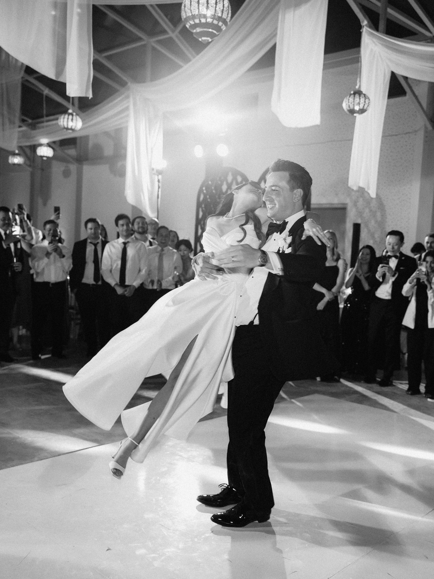 Black and white photo of groom lifting bride during first dance at elegant wedding reception.