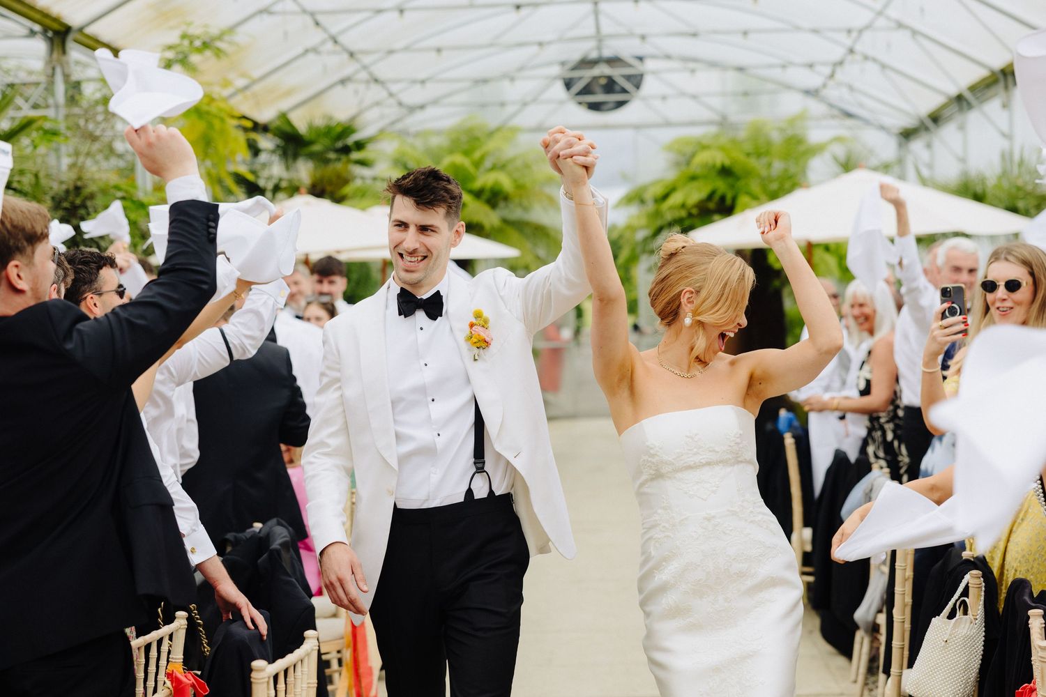 Bride and groom celebrate recessional in greenhouse wedding venue with guests waving white handkerchiefs.