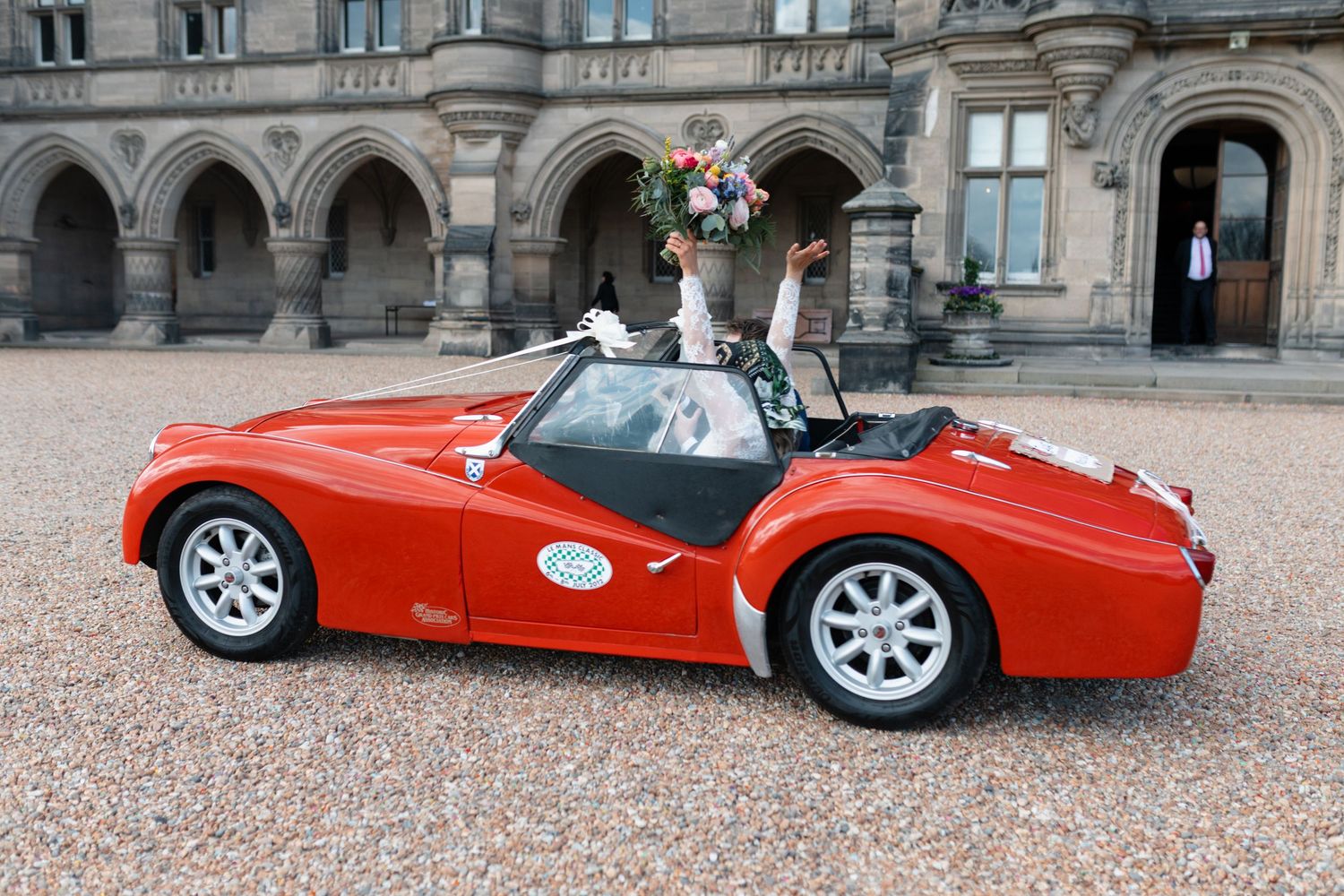 Red vintage convertible sports car with wedding flowers, parked outside a grand stone building with a bride holding a bouquet.