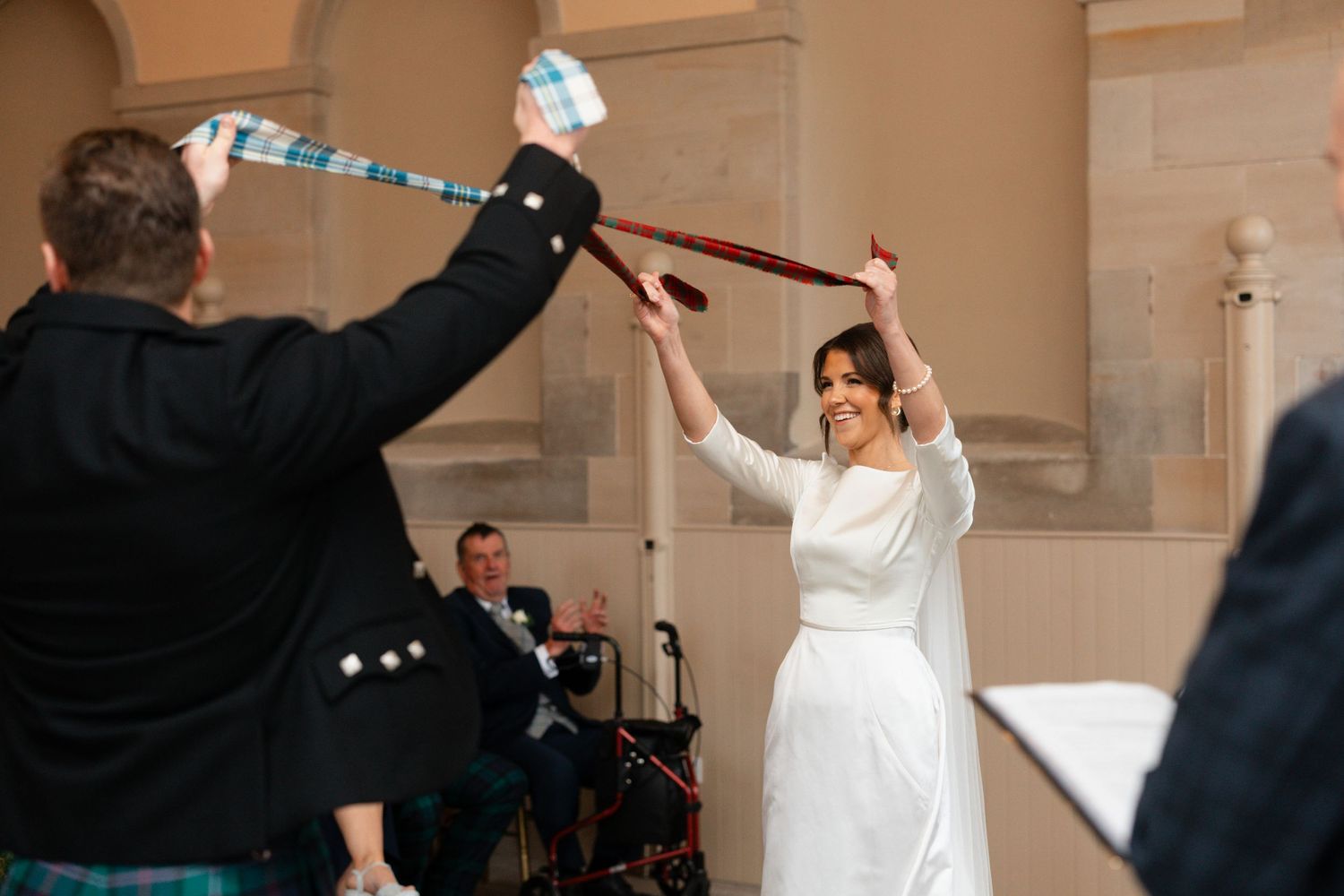 Bride and groom perform a traditional Scottish handfasting ceremony, raising tartan ribbons in an elegant stone venue.