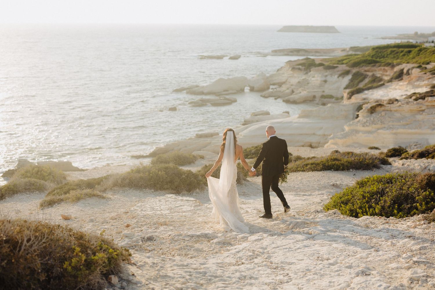 Bride and groom walking hand in hand along scenic coastal cliffs, brides veil flowing in breeze.