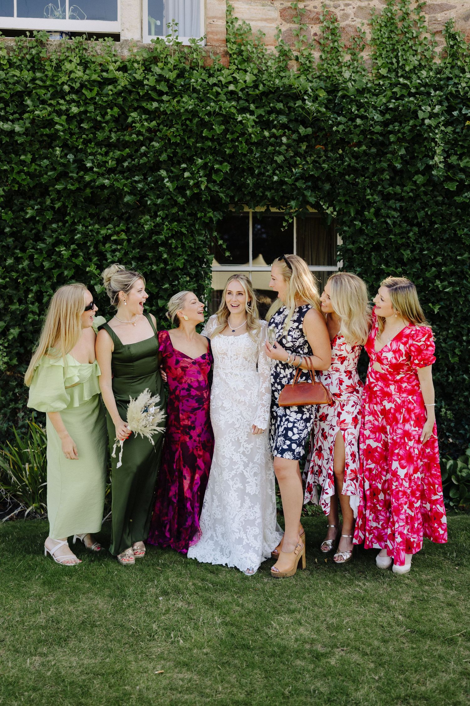 Bride in white lace gown poses with bridesmaids in colorful dresses on green lawn before ivy-covered wall.