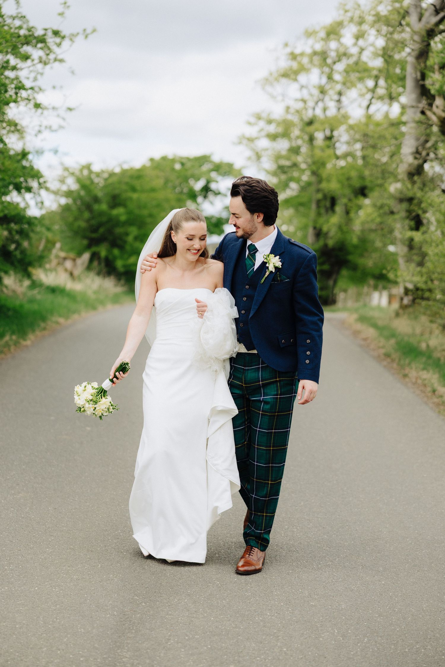 A bride and groom walk together on a country road, she in a white gown with veil, he in a navy jacket and tartan kilt.