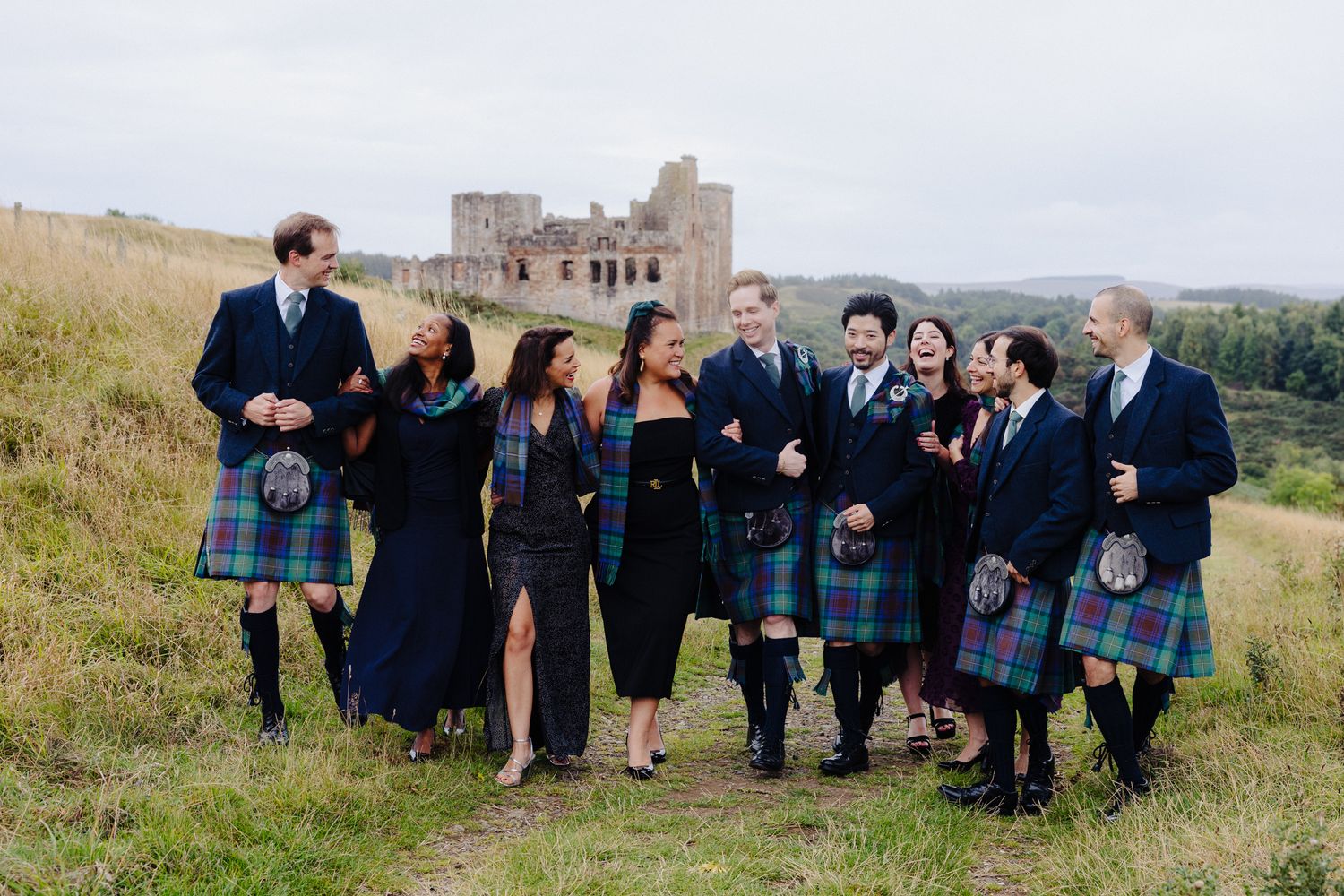 Wedding party in kilts and dark formal wear laughing together before a Scottish castle ruin backdrop.