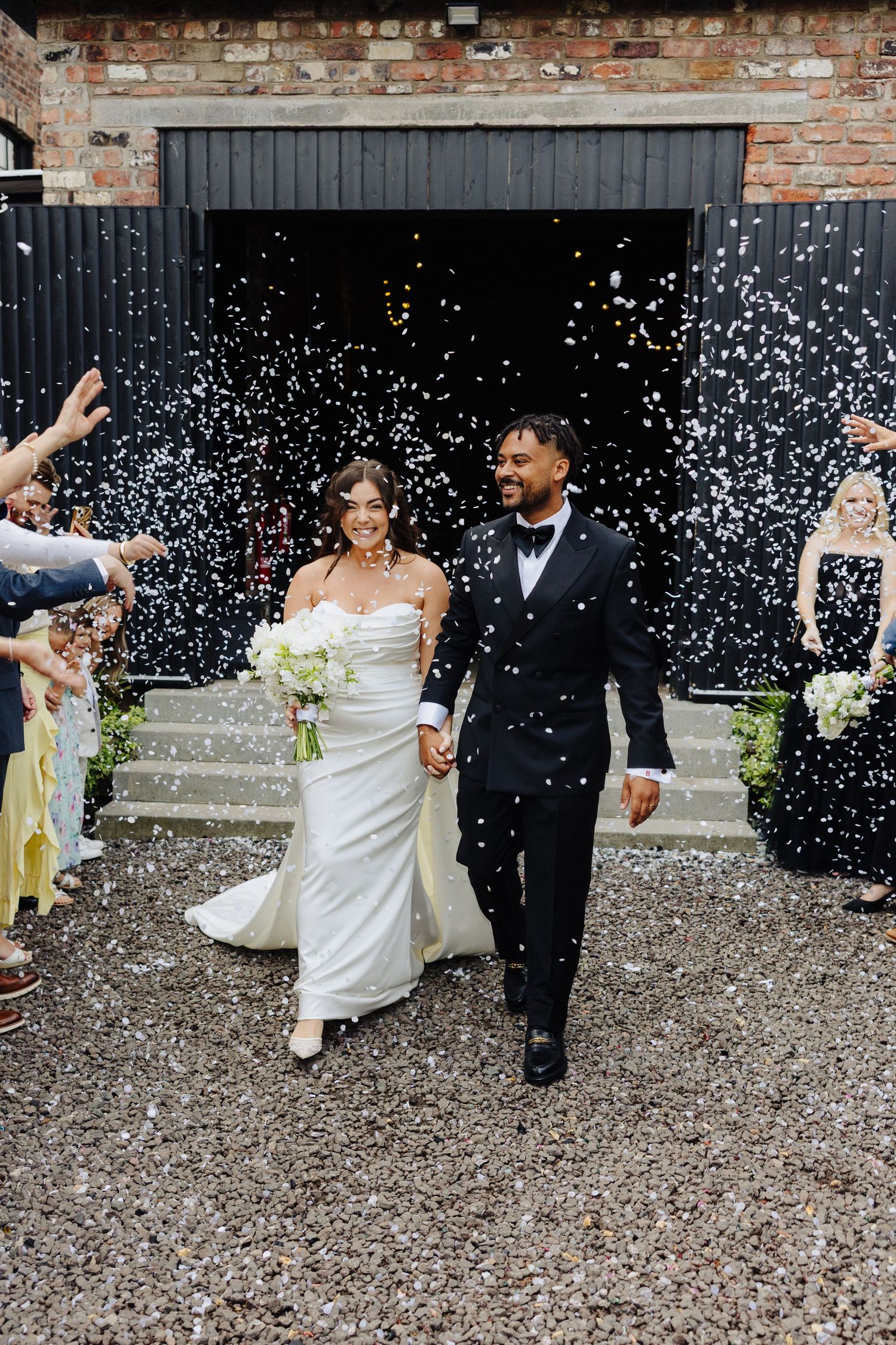 Happy newlyweds walking through a shower of white confetti outside a rustic barn venue.