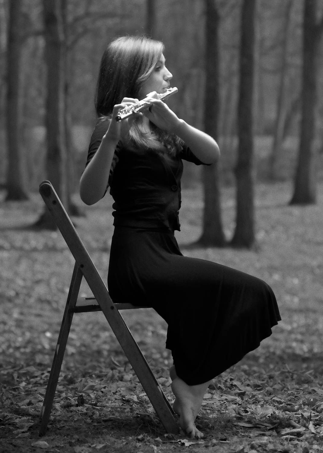 Creative black and white portrait of young woman with dark hair sitting on a chair playing the flute outside in the woods