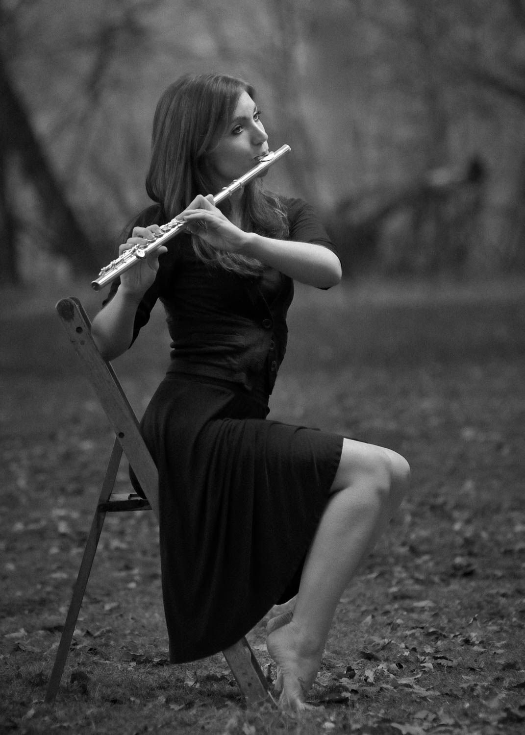 Creative black and white portrait of young woman with dark hair sitting on a chair playing the flute outside in the woods