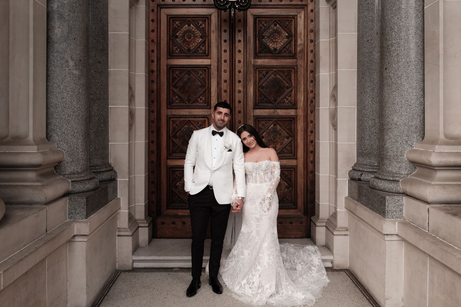 Bride and groom standing before ornate wooden doors of a classical stone building on their wedding day.