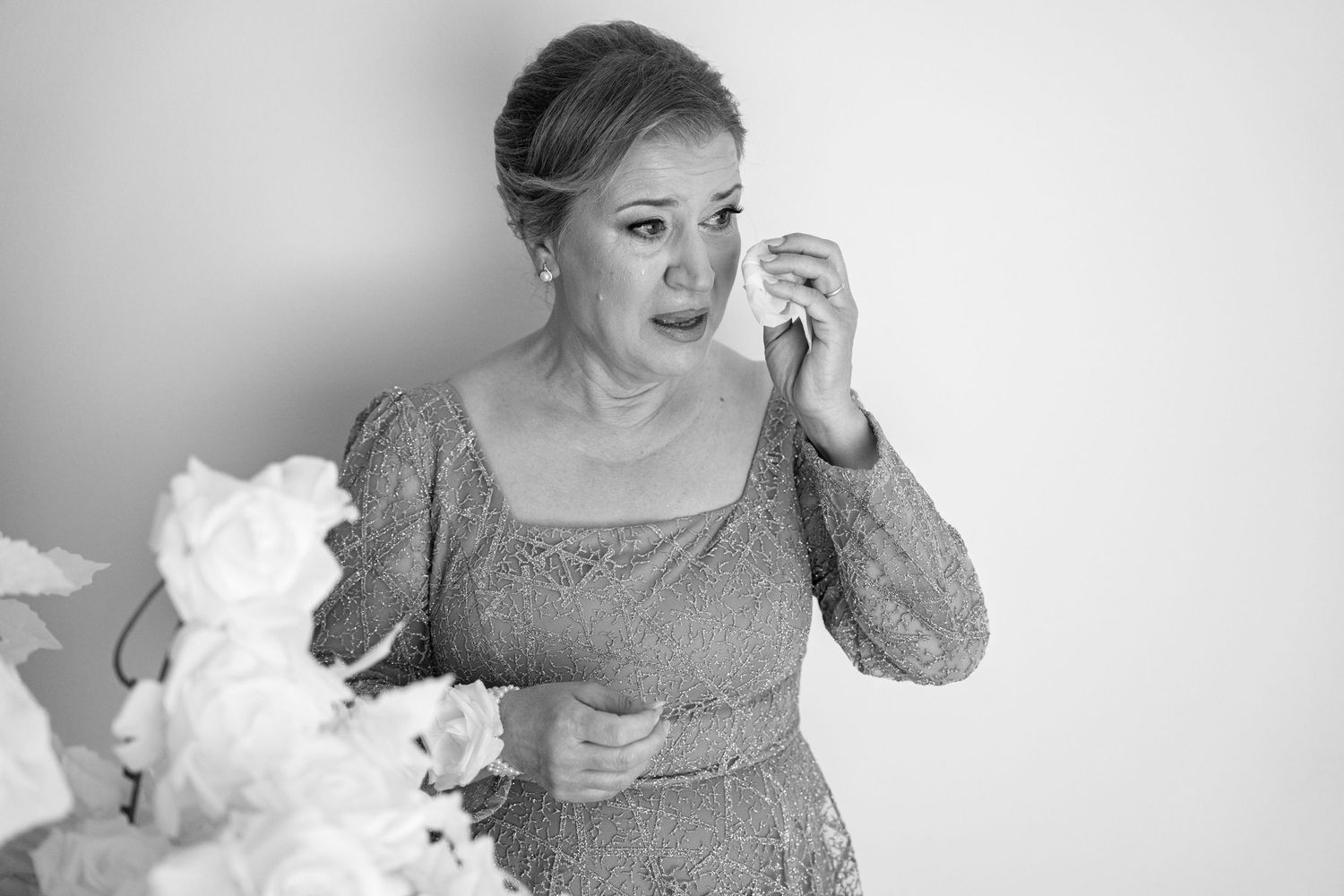 Emotional woman in lace dress wiping tears beside white flowers in black and white wedding photo.