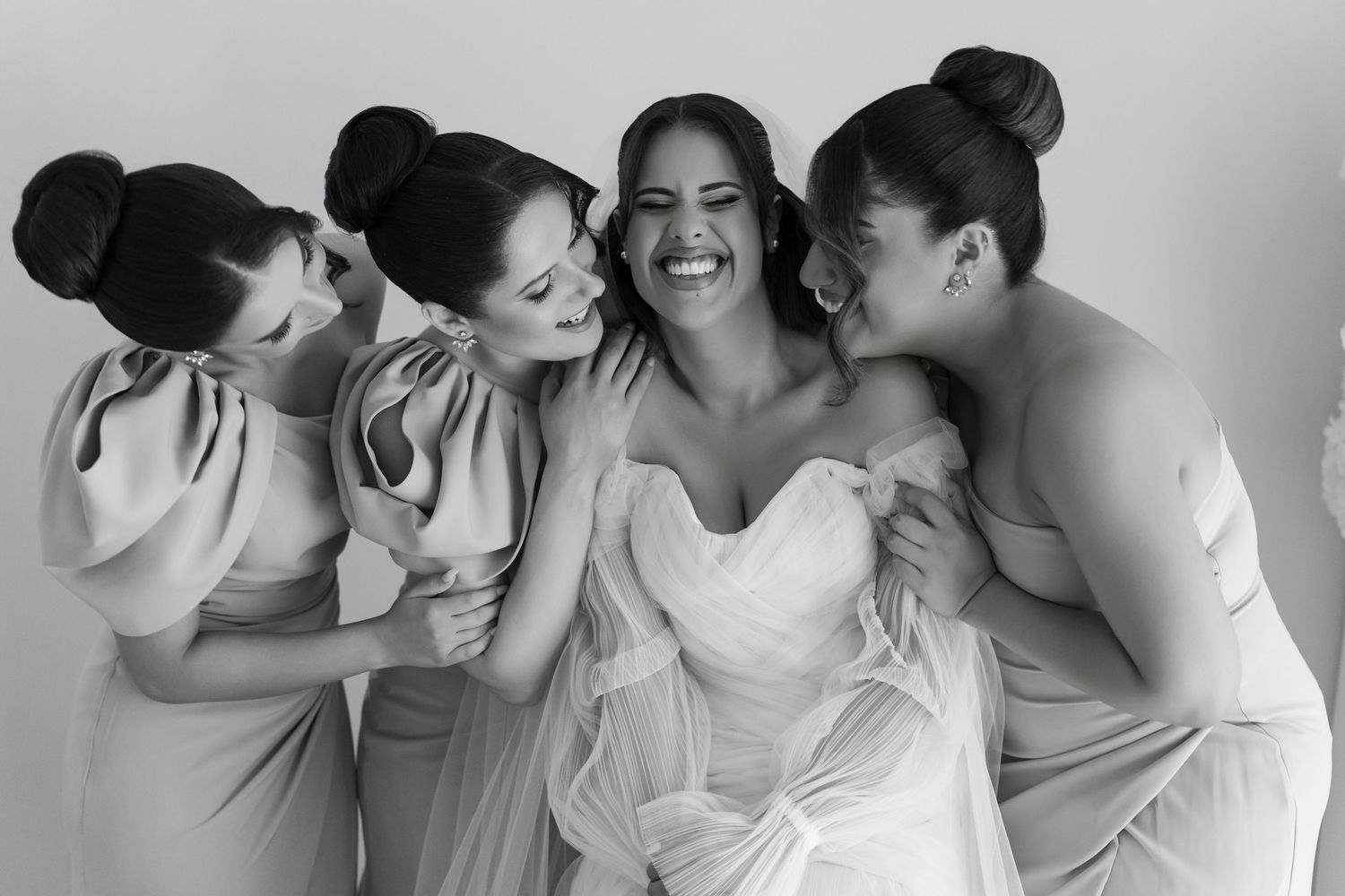 Three bridesmaids in satin dresses embracing laughing bride in black and white wedding portrait.