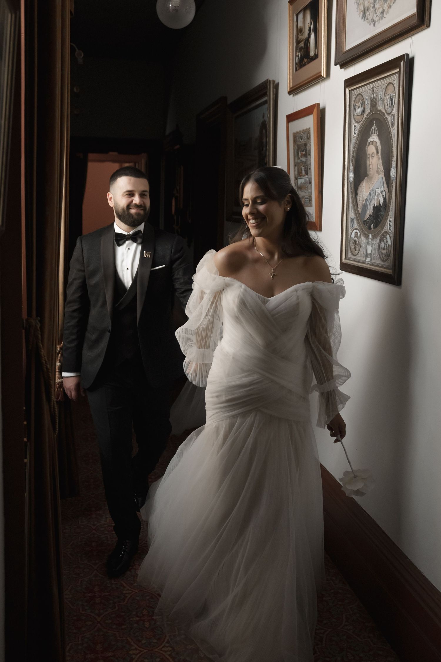 Bride in off-shoulder tulle gown walks with groom in tuxedo through hallway lined with portraits.