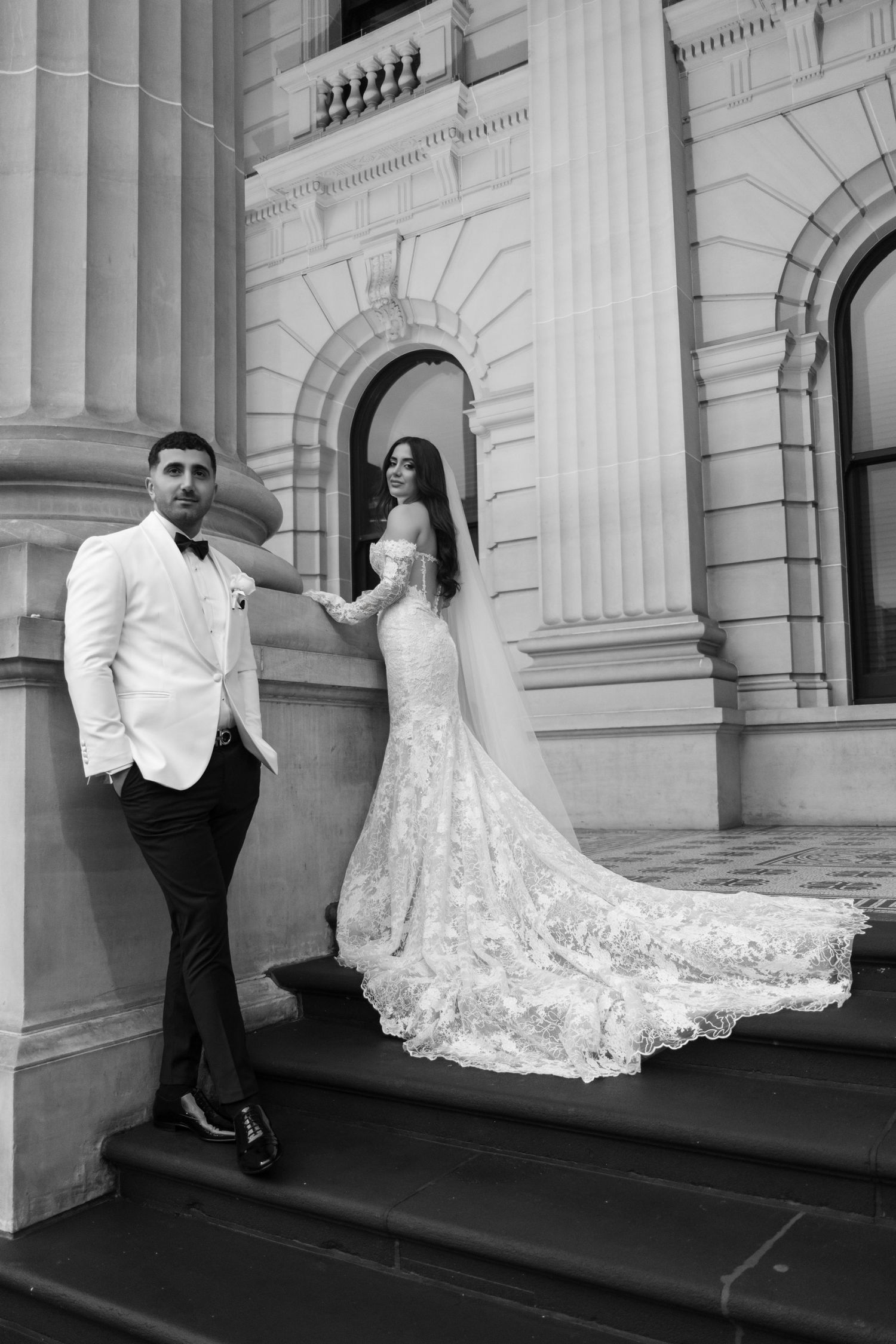 Black and white photo of a bride and groom posing on steps of a grand classical building.