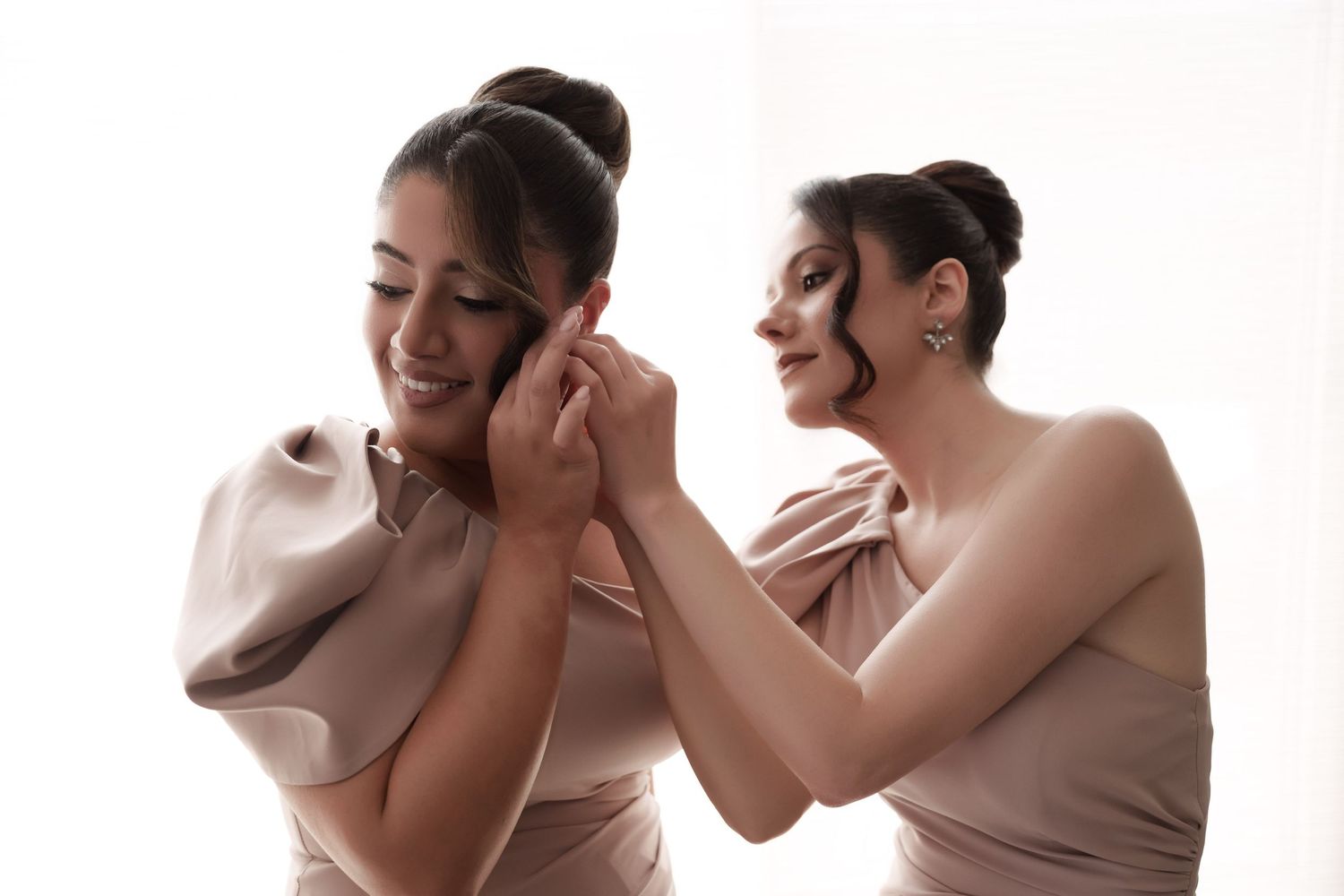 Two bridesmaids in blush satin dresses laughing while helping each other with earrings.